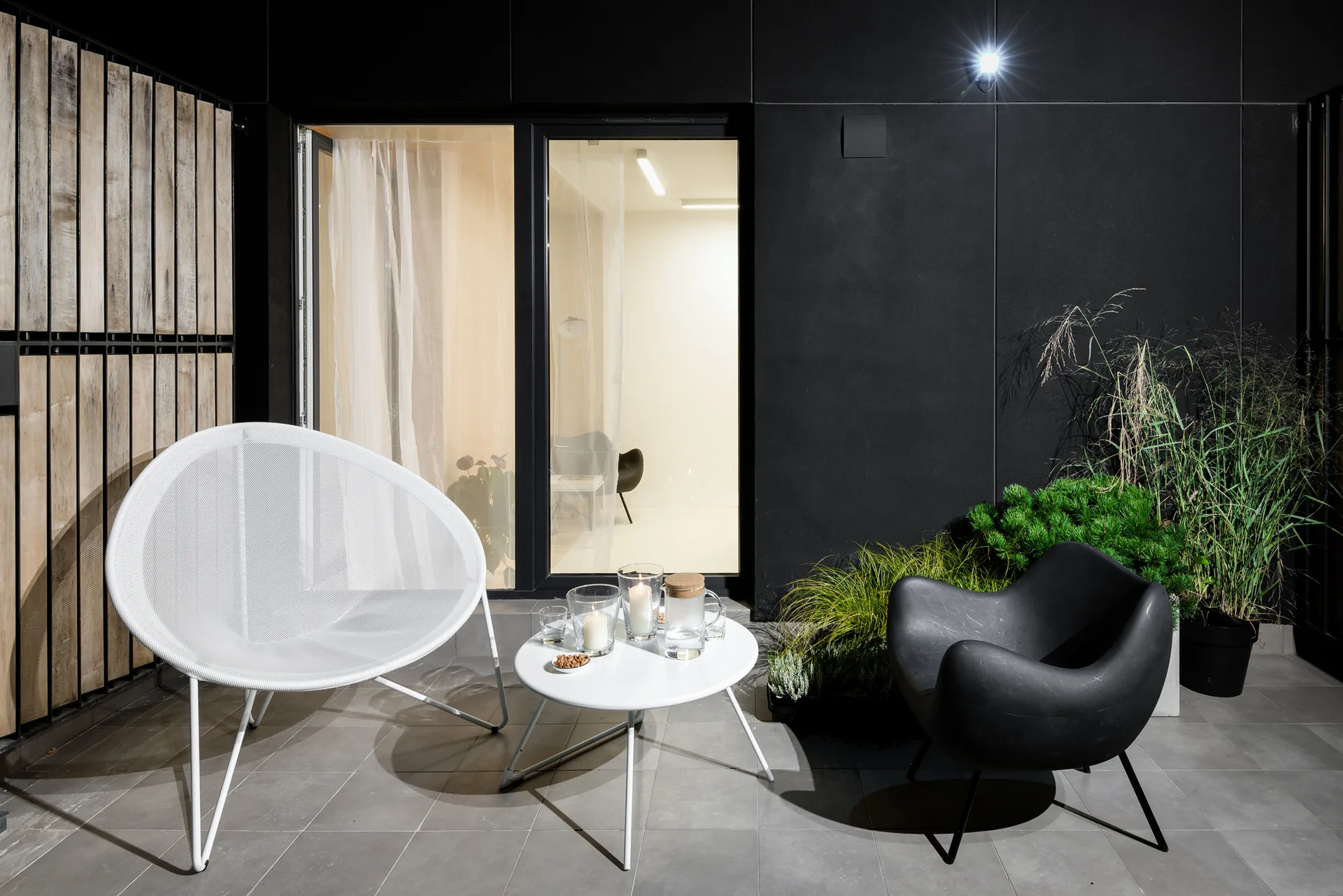 A modern night terrace features a white mesh armchair and a black molded plastic chair around a small white table with candles against a dark wall.