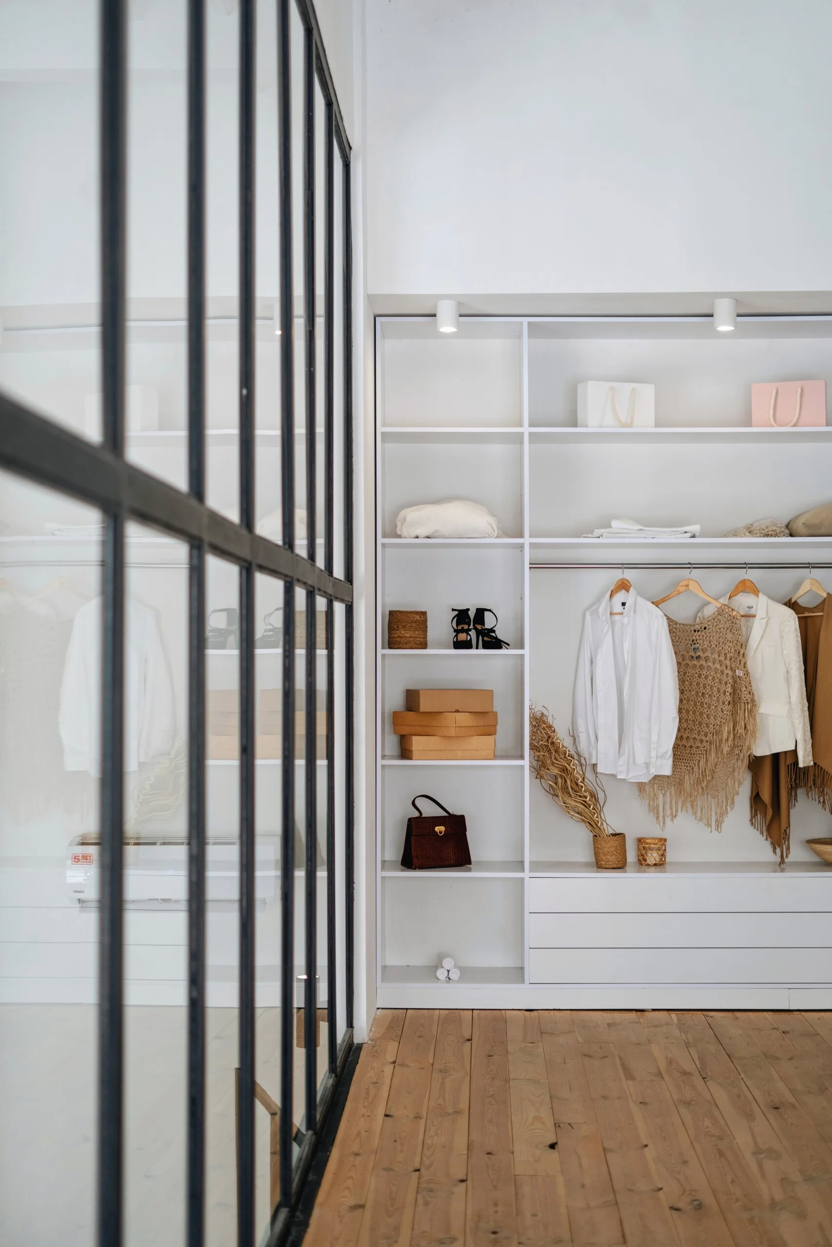 A minimalist walk-in closet featuring white open shelving, hanging neutral clothing, and a black-framed glass partition by light wood flooring.