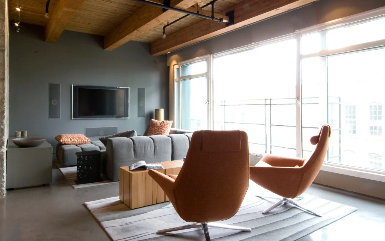 An industrial living room featuring a grey sectional sofa, two orange swivel armchairs, a wood-beamed ceiling, and large windows.