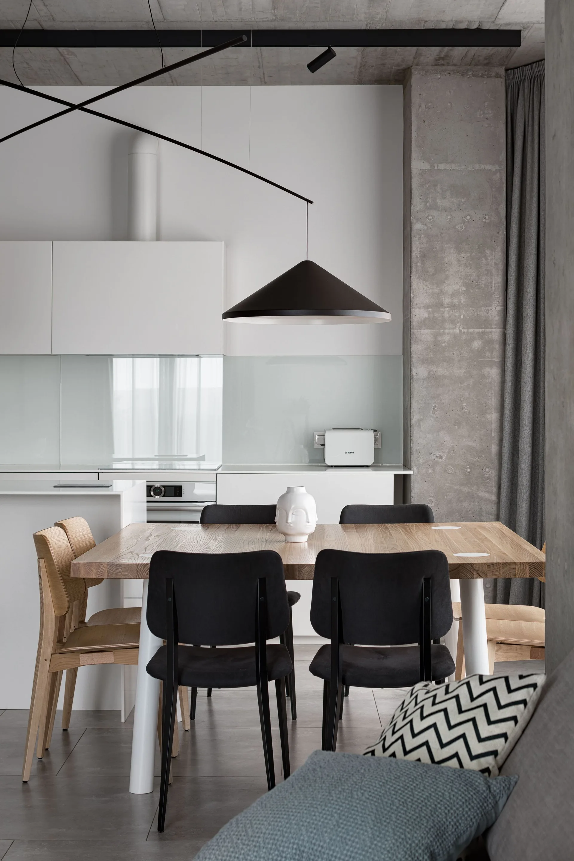 A modern dining area features a light wood table with black and wooden chairs, an overhead black pendant light, and an exposed concrete column.