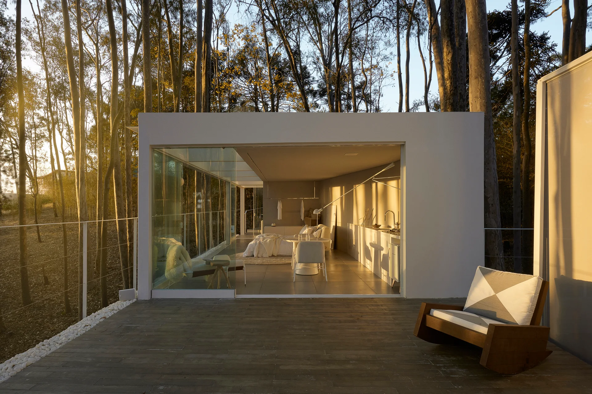 A modern glass-walled studio cabin nestled in a forest opens onto a wooden deck featuring a single wooden rocking chair during golden hour.