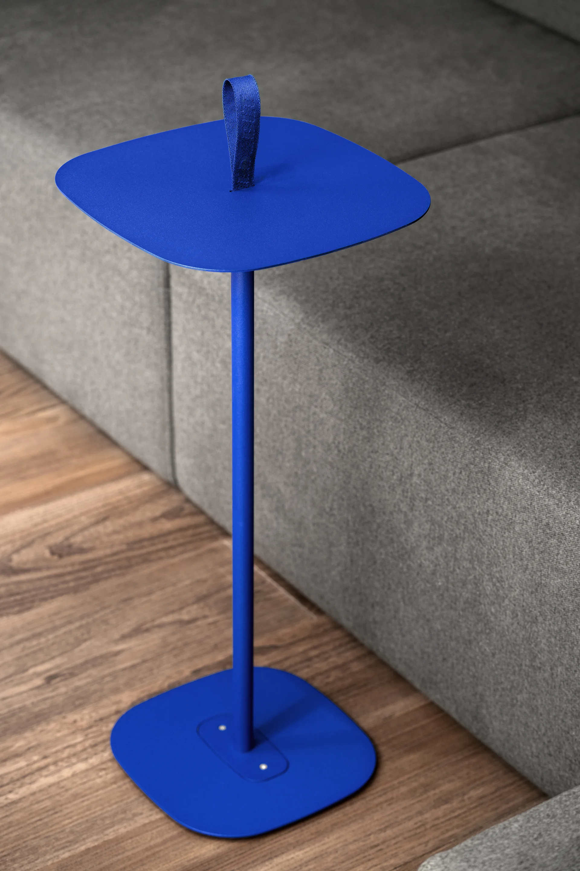 A bright blue metal pedestal side table with a fabric strap handle stands on a wood floor next to a grey sofa.