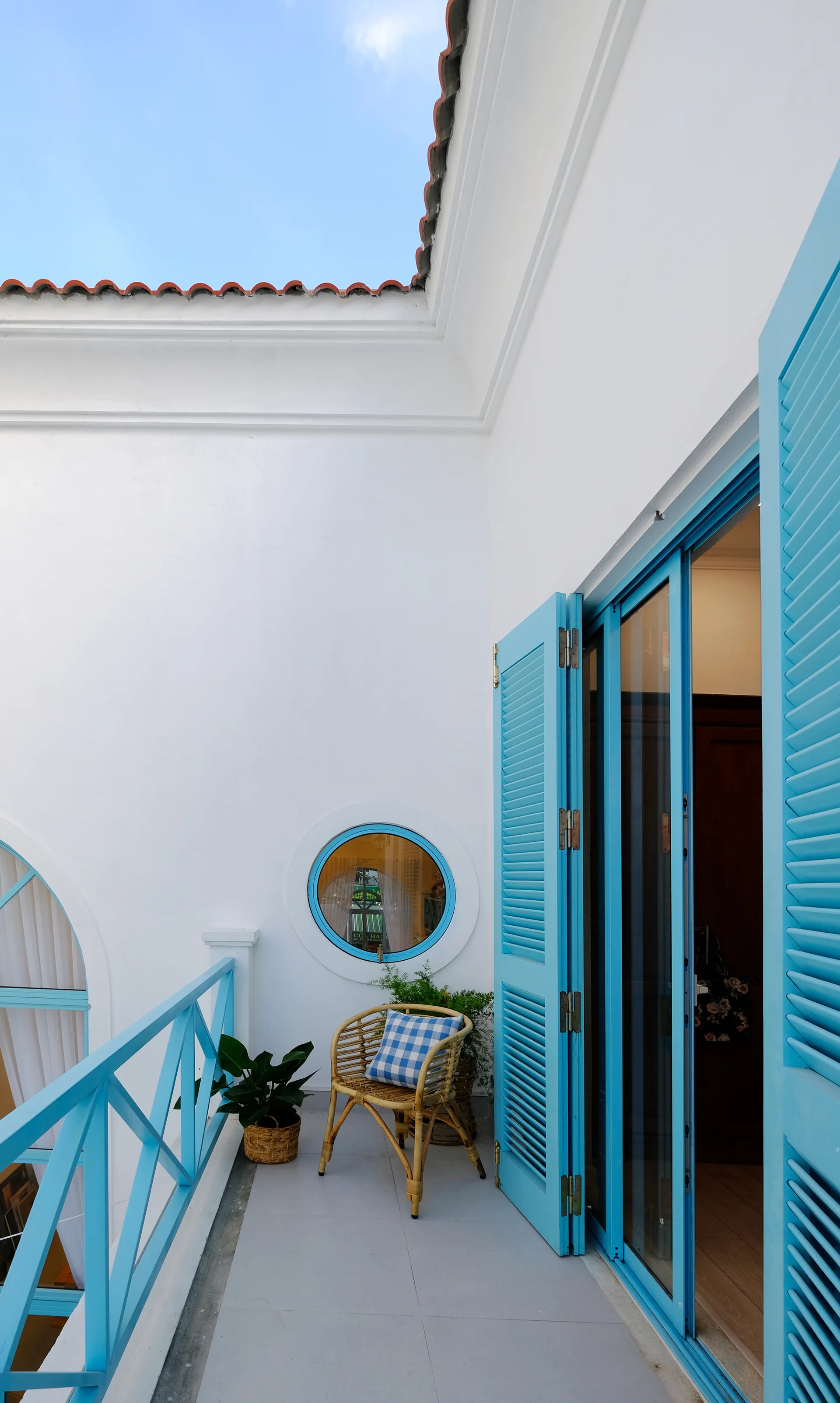A white outdoor balcony features bright blue louvered shutters, a matching wooden railing, a circular porthole window, and a wicker chair.