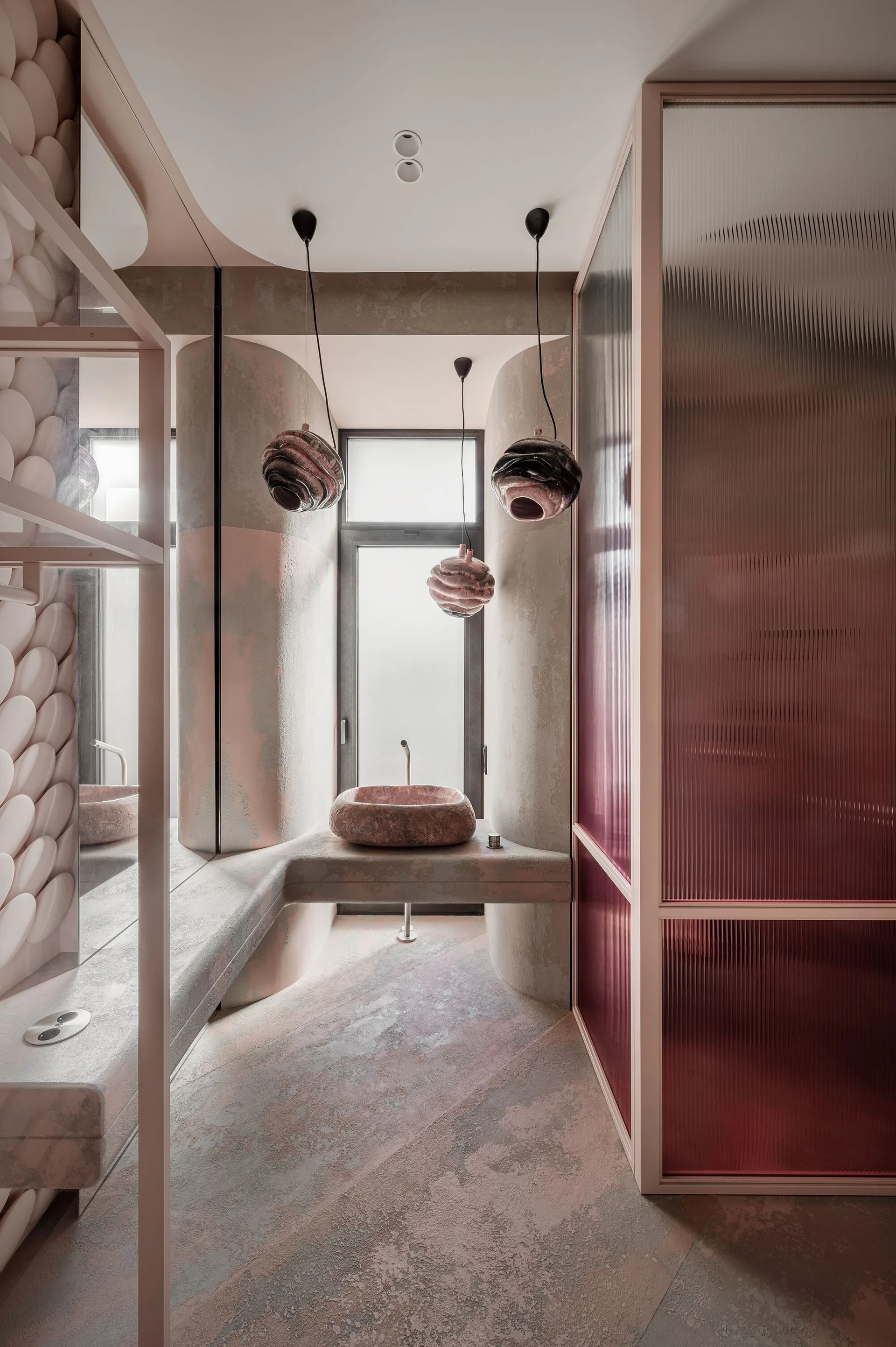 A modern powder room features a stone vessel sink on a floating concrete vanity, sculptural pendant lights, and pink-tinted fluted glass panels.