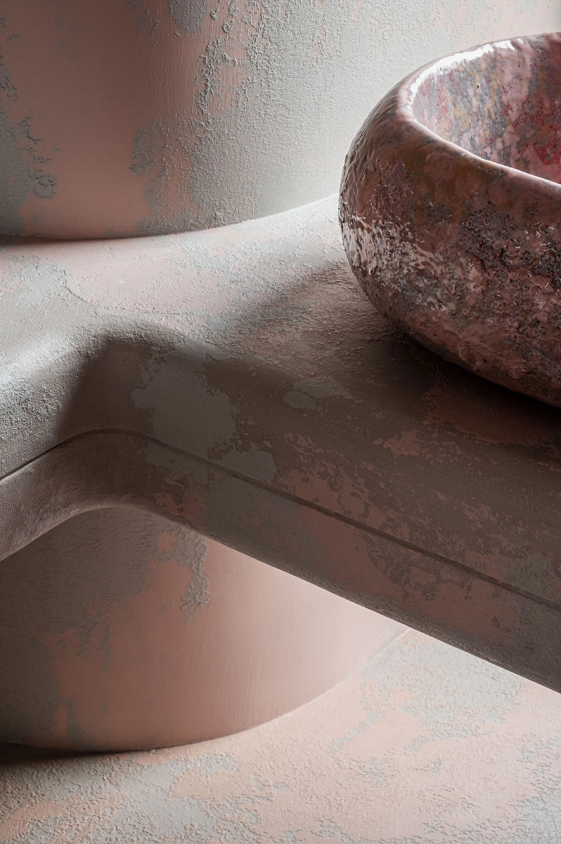 A close-up of a sculptural, weathered pink stone basin sitting on a curved, matching textured pink plaster pedestal.