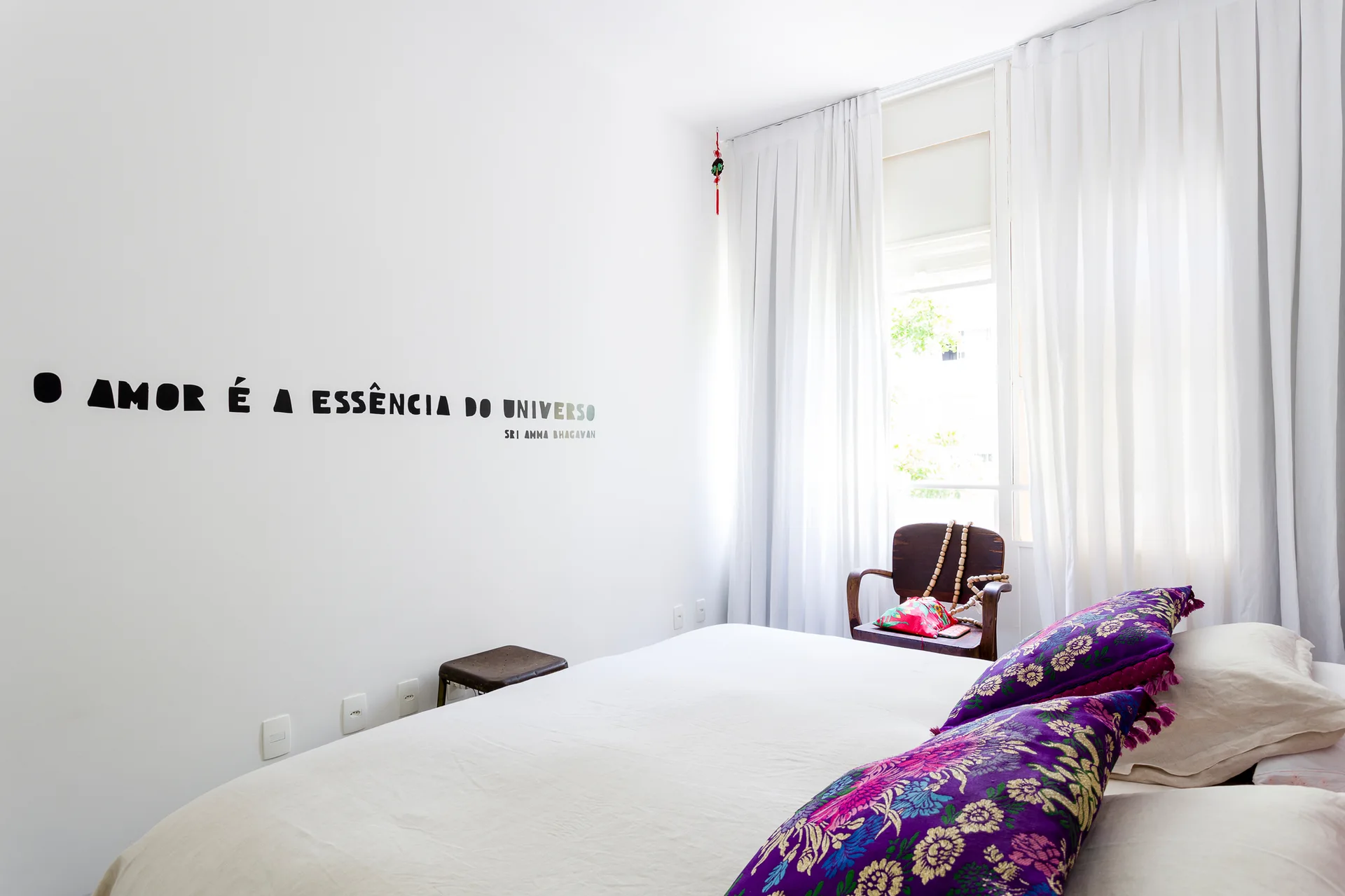 A minimalist white bedroom featuring a black wall decal, white bedding accented by purple floral pillows, and a wooden armchair by the window.