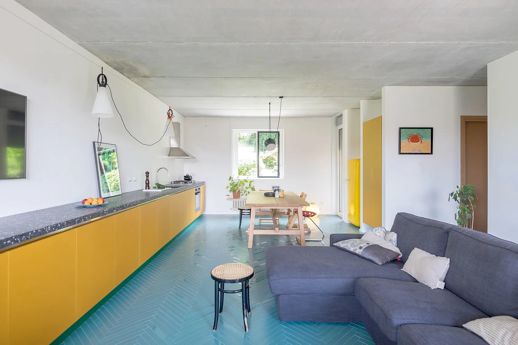 An open-plan kitchen and living space featuring bright yellow cabinets, a glossy teal herringbone tile floor, a wooden dining table, and a grey sofa.
