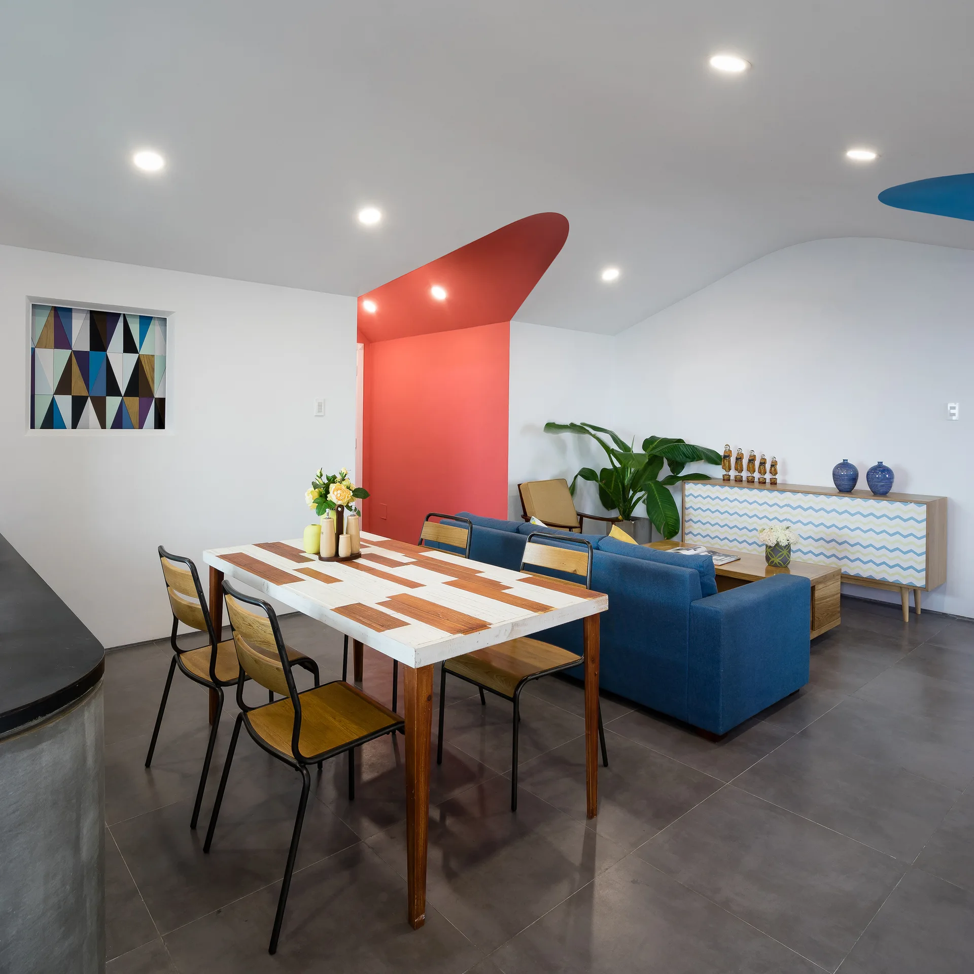 A modern open-plan dining and living area featuring a wood dining table, a blue sofa, and a curved white ceiling with a bold red cutout section.