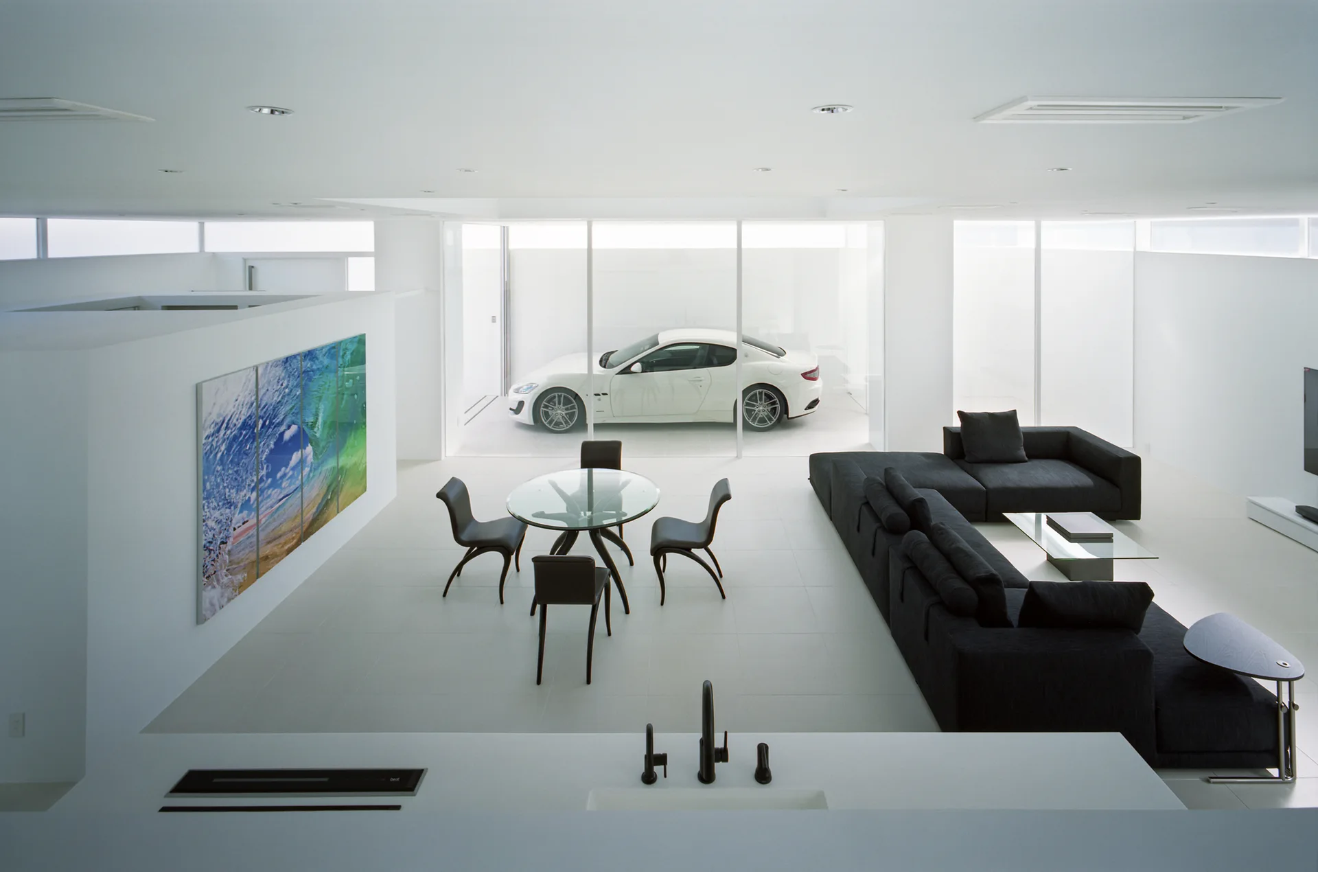 A minimalist white living room features a black sectional sofa, a glass dining table, and a white sports car visible behind a glass wall.