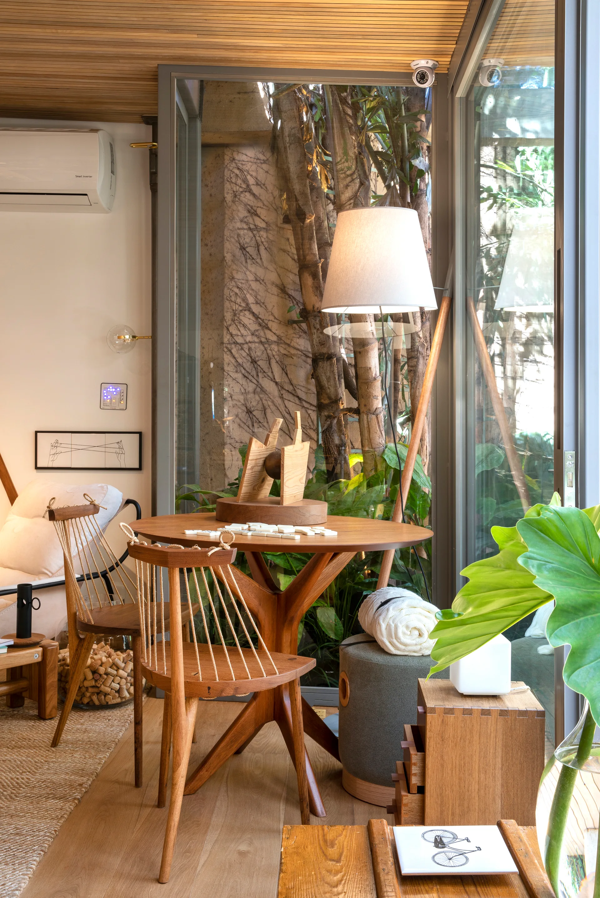 A round wooden dining table with spindle chairs stands next to floor-to-ceiling windows showing a lush green courtyard.