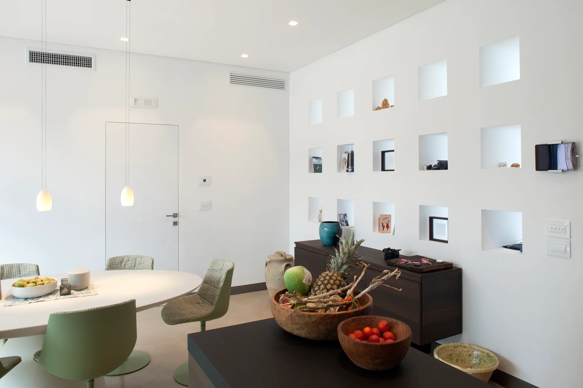 A modern dining room with an oval white table, green chairs, a dark wood sideboard, and a white wall featuring a grid of square recessed niches.