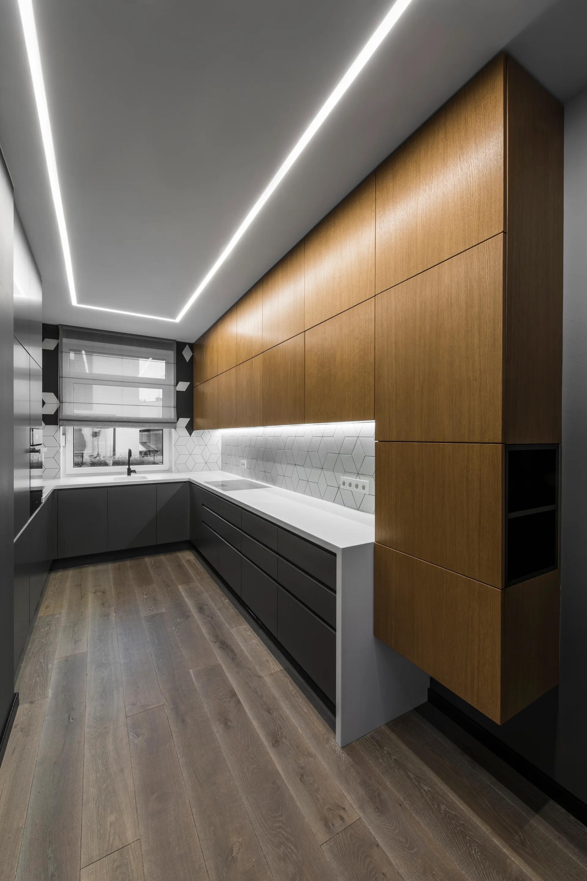 A modern galley kitchen features warm wood upper cabinets, matte charcoal lower cabinets, and a white geometric tile backsplash.