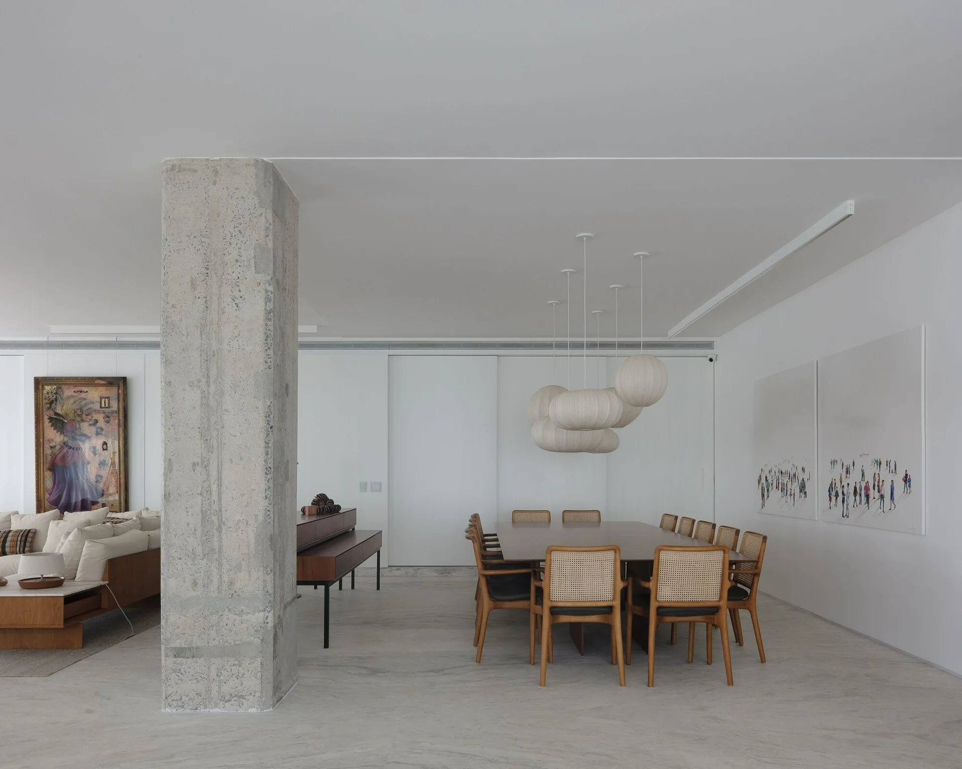 A minimalist dining room featuring a long wooden table with cane-back chairs, a paper pendant light cluster, and a large concrete column.