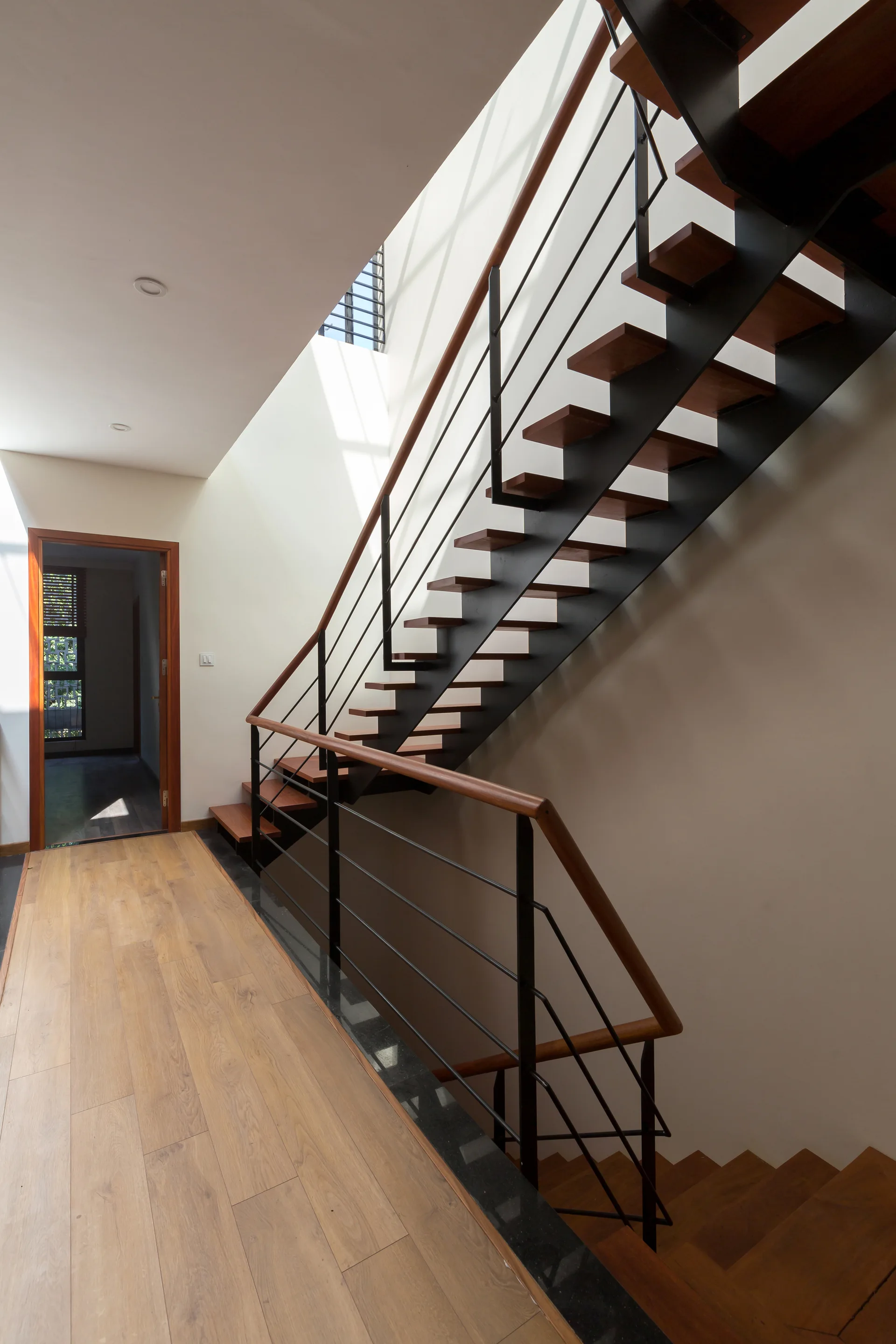A modern open-riser wood staircase with black metal railings stands in a bright hallway with light-colored wood flooring and clean white walls.