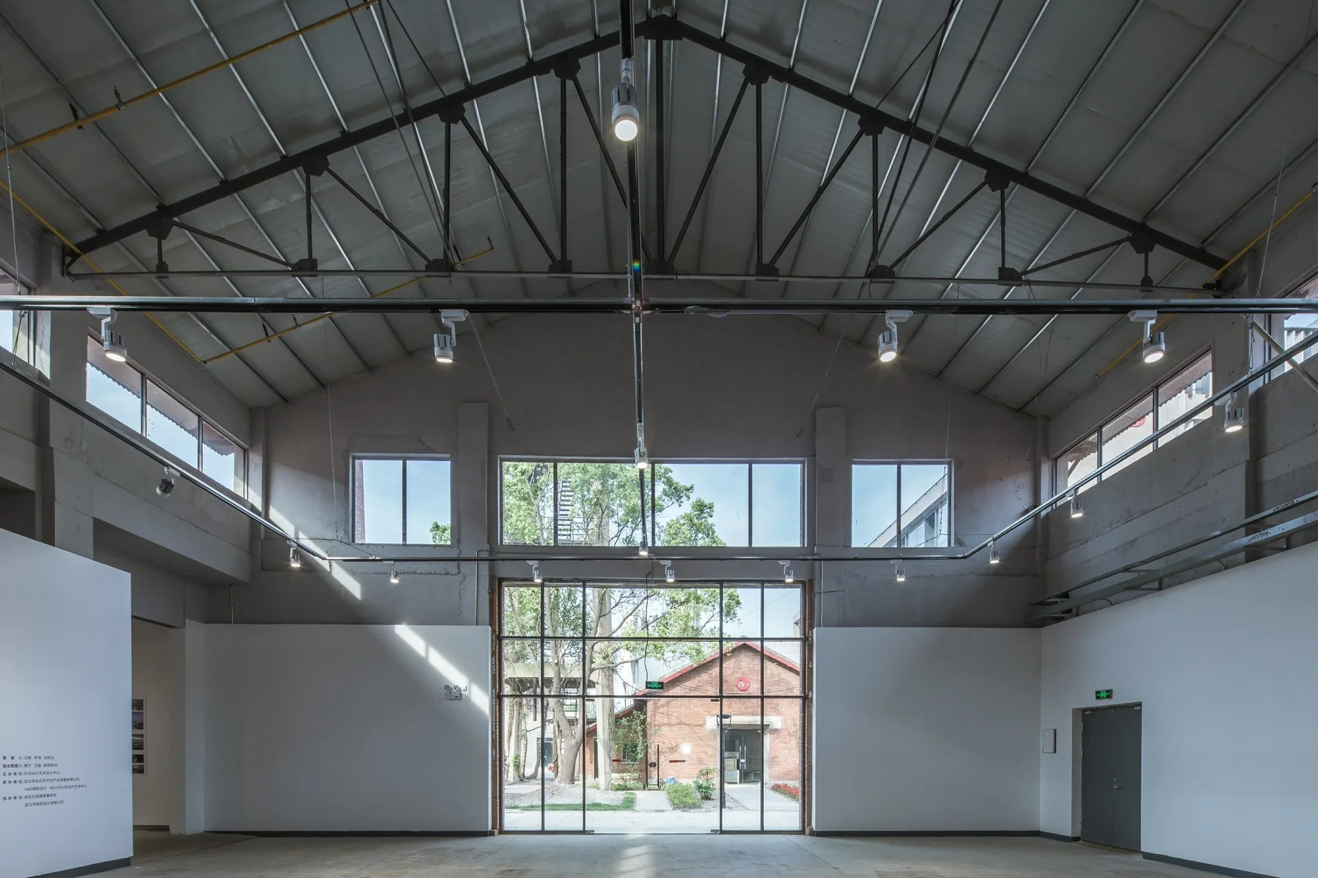 A spacious industrial gallery interior features a gabled ceiling with exposed steel trusses, white display walls, and large steel-framed glass doors.