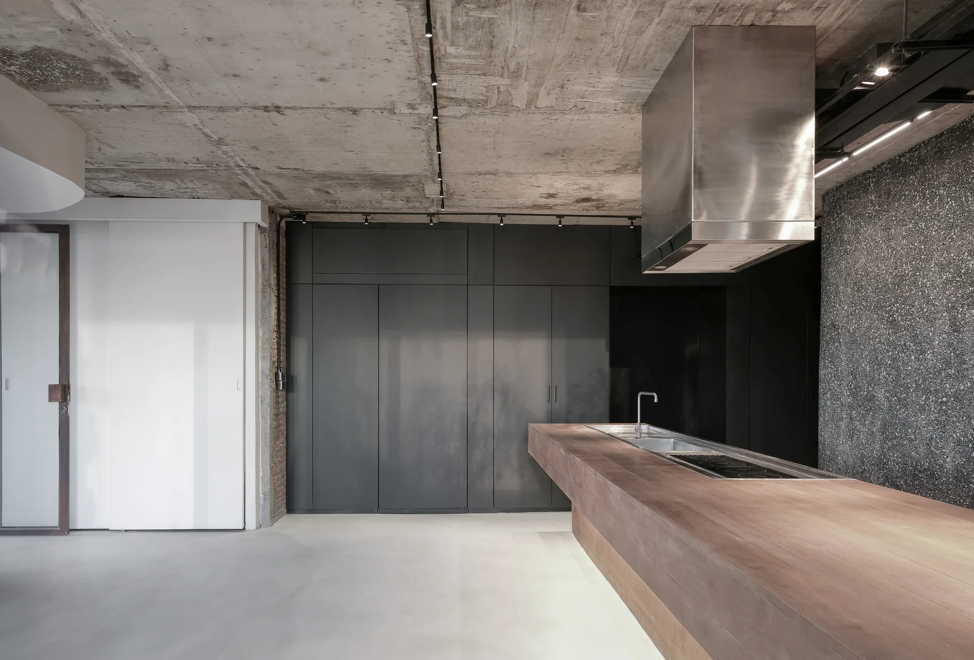 A minimalist kitchen features a long wooden island with an integrated sink and cooktop beneath an exposed concrete ceiling.