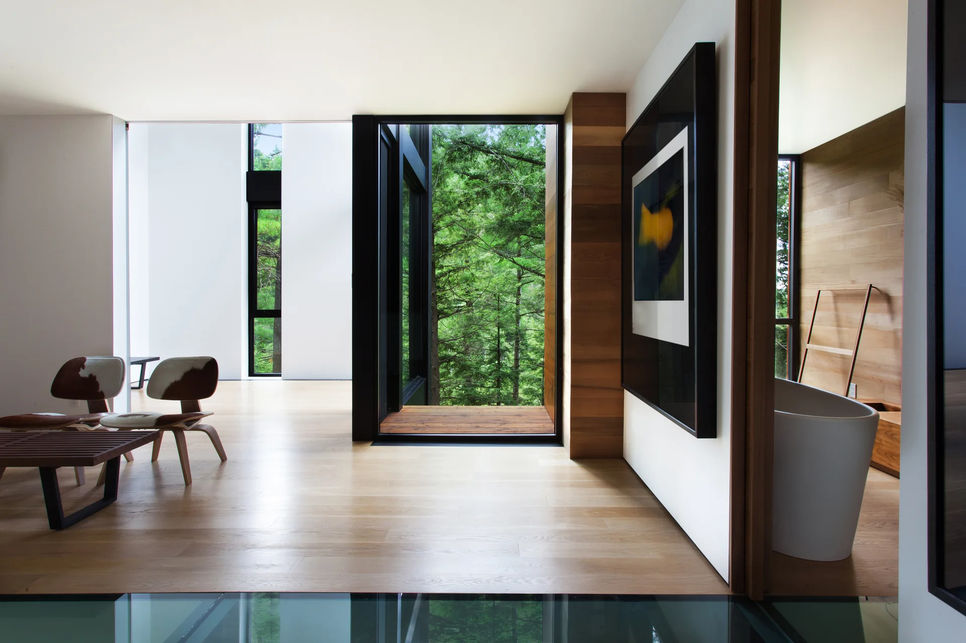 A modern interior with light wood flooring, mid-century chairs, a glass floor section, and a large window framing green forest trees.