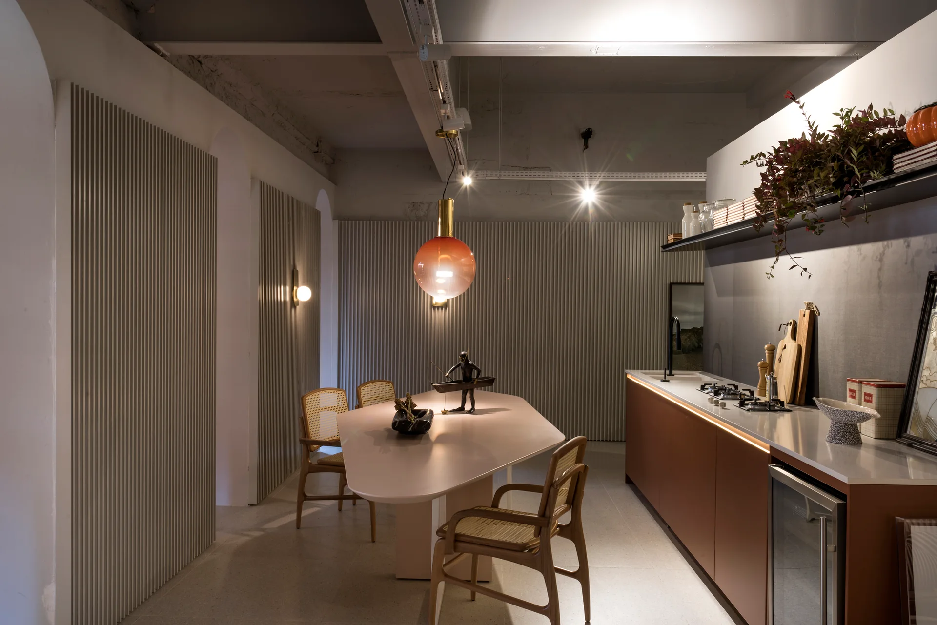 A modern kitchen features brown lower cabinets with under-counter lighting, a sculptural light beige dining table, rattan chairs, and vertical slatted walls.