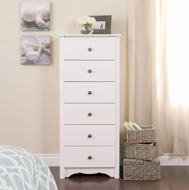 A tall white six-drawer dresser stands on dark wood flooring next to a stack of three grey woven baskets and a light-filtering window curtain.