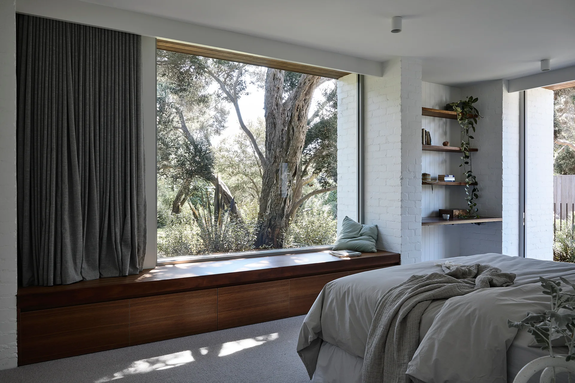 A minimalist bedroom featuring a large picture window, a built-in wood window bench, and open wooden shelving against a painted white brick wall.