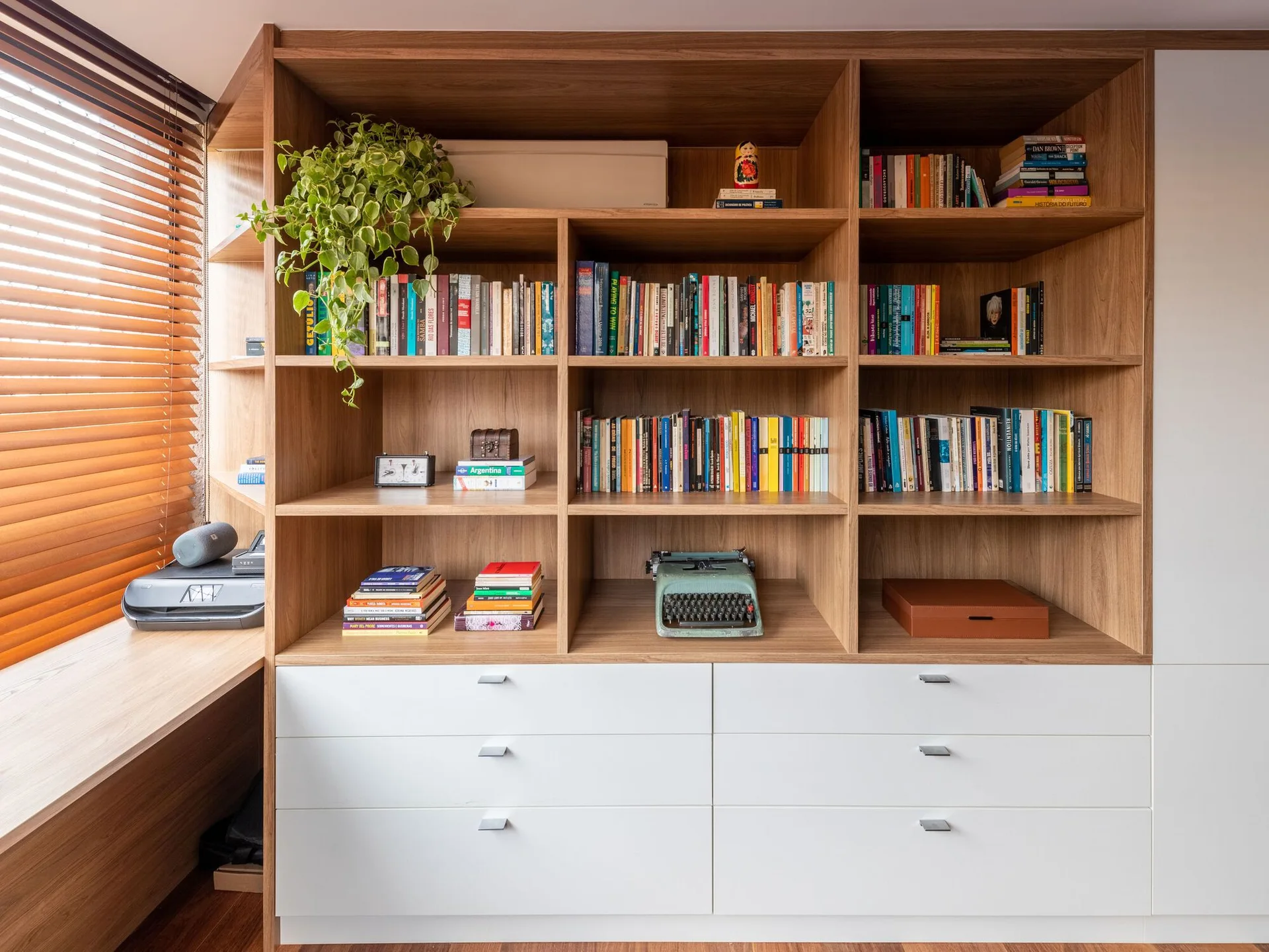 A built-in wooden bookshelf with open compartments holding books sits above white drawers next to an integrated desk and wooden window blinds.