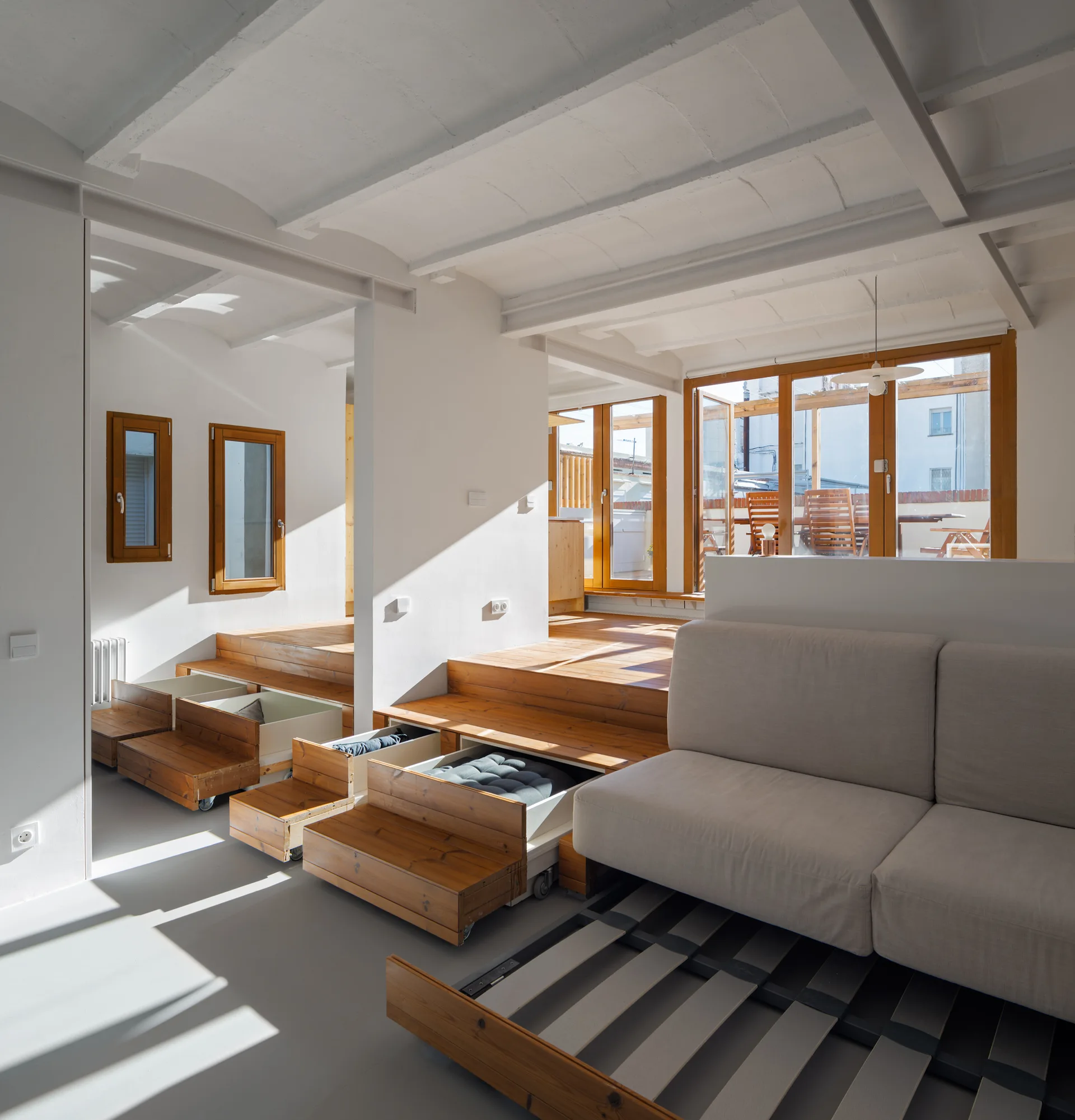 A modern multi-level living room featuring natural wood tiered floors with pull-out storage drawers, a light gray sofa, and wooden window frames.