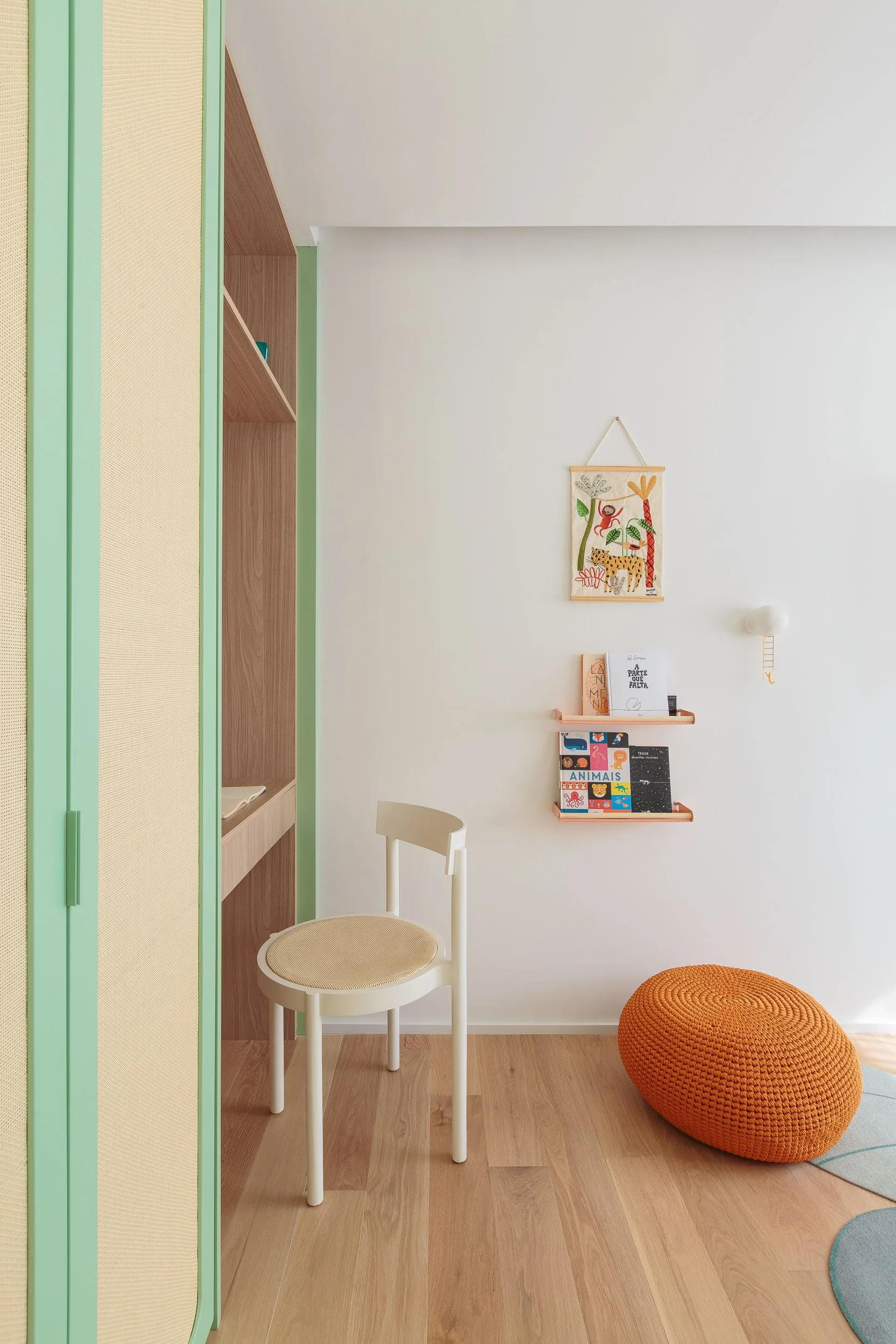 A children's study area features a mint green and cane built-in cabinet, a white wooden chair, a wood-toned desk, and an orange knit pouf on wood flooring.