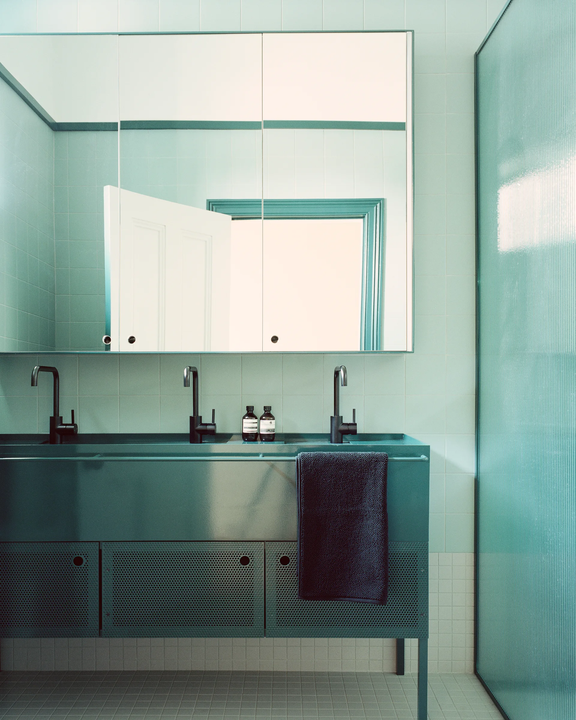 A modern bathroom features a long teal metal vanity with perforated doors and black faucets set against light blue tiled walls.
