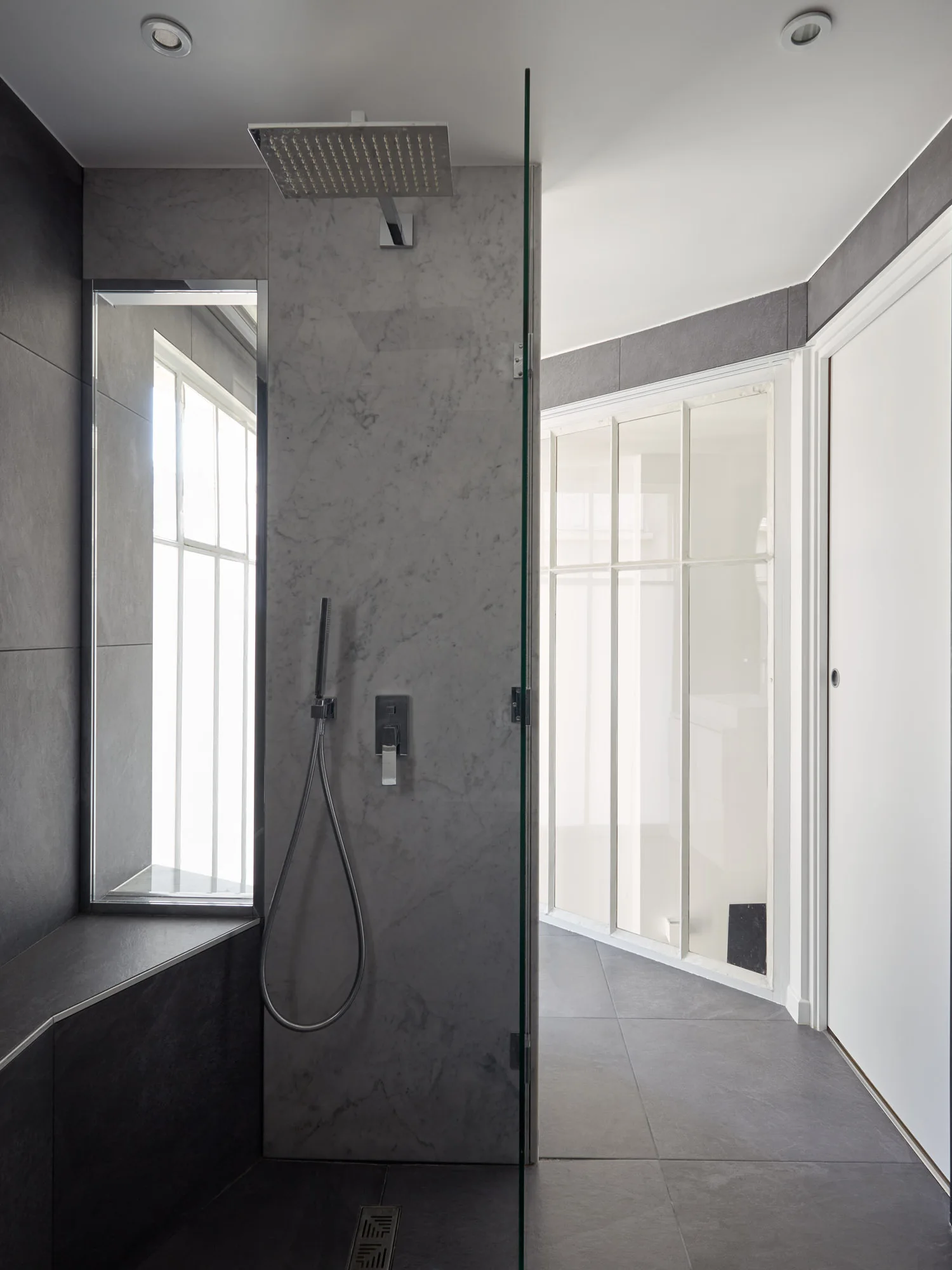A modern walk-in shower features dark gray stone-look tiles, a light gray marble accent panel, a rainfall showerhead, and a frameless glass partition.