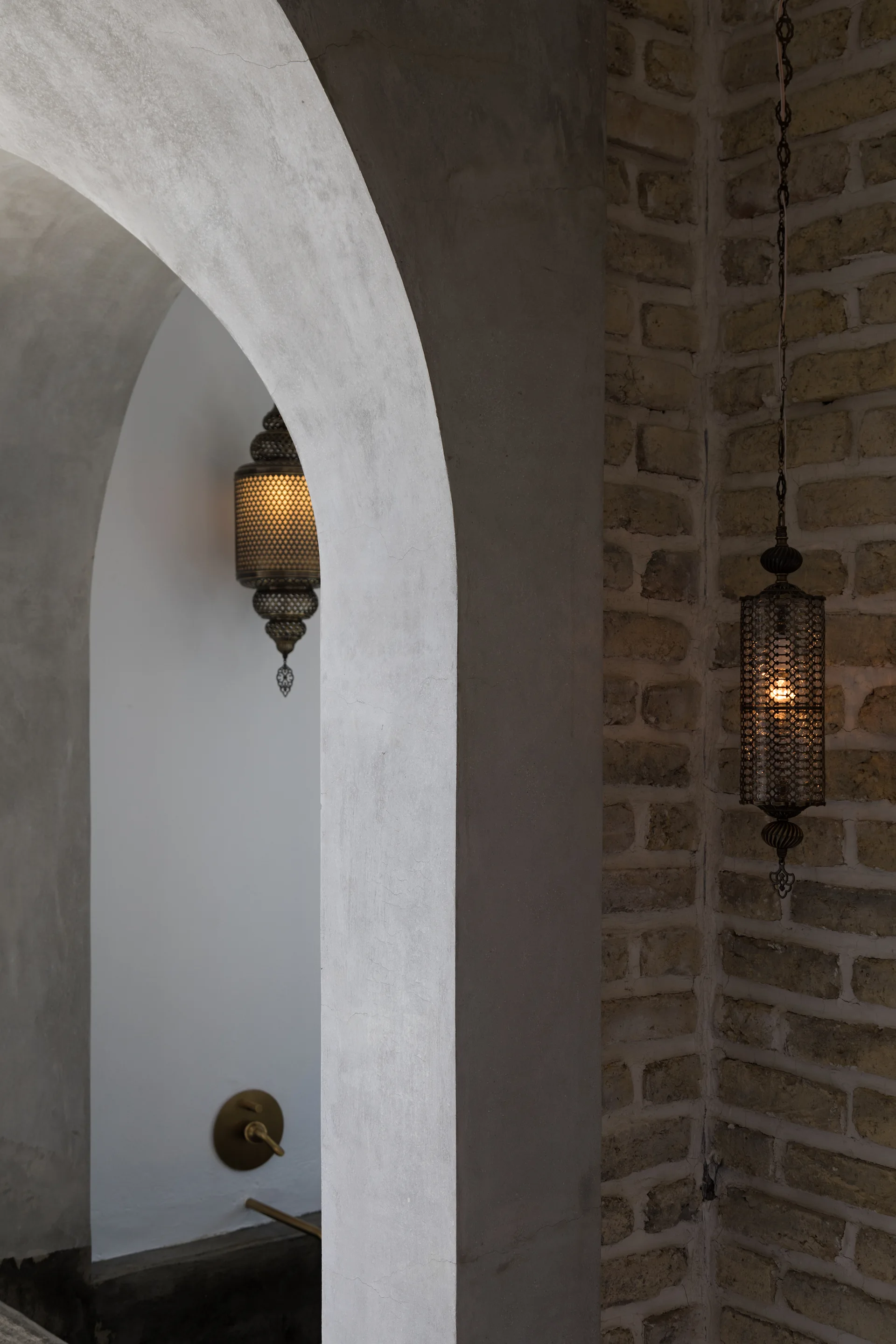 A view through a smooth plaster archway shows an exposed brick wall decorated with a hanging metal lantern emitting a warm glow.