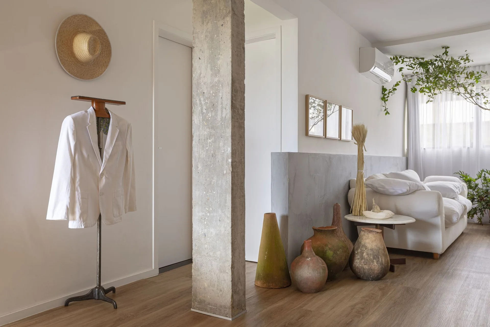 A bright living space features an exposed concrete column, wood floors, and a collection of earthy pottery vessels next to a white sofa.