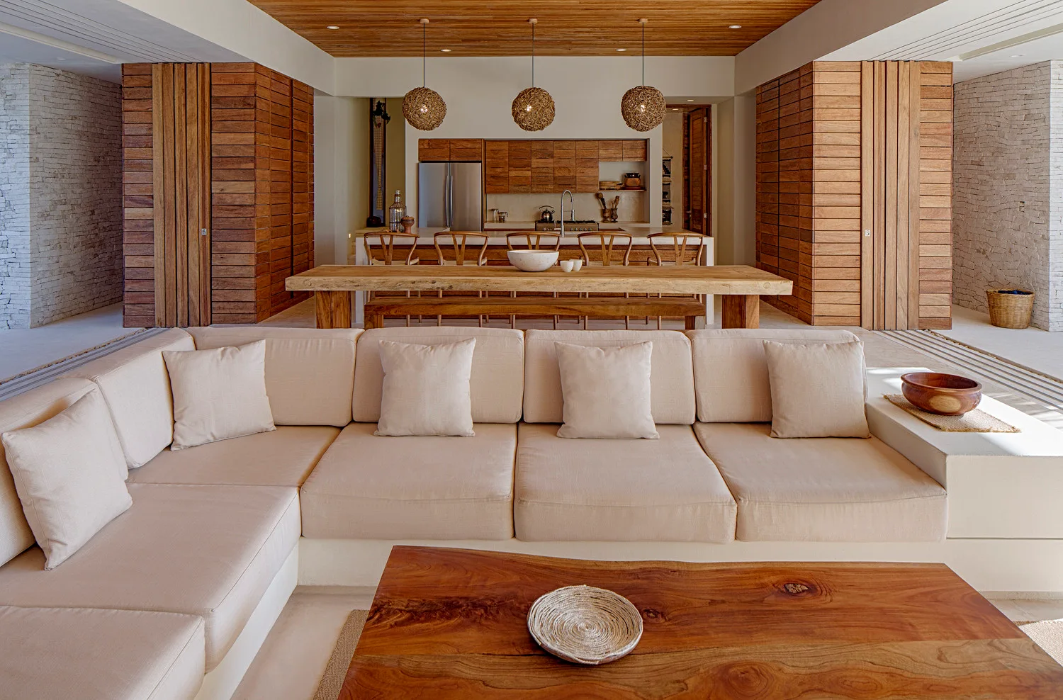 An open-plan living and dining space features a low cream sectional sofa, a live-edge wood dining table, and a horizontal wood slat ceiling.