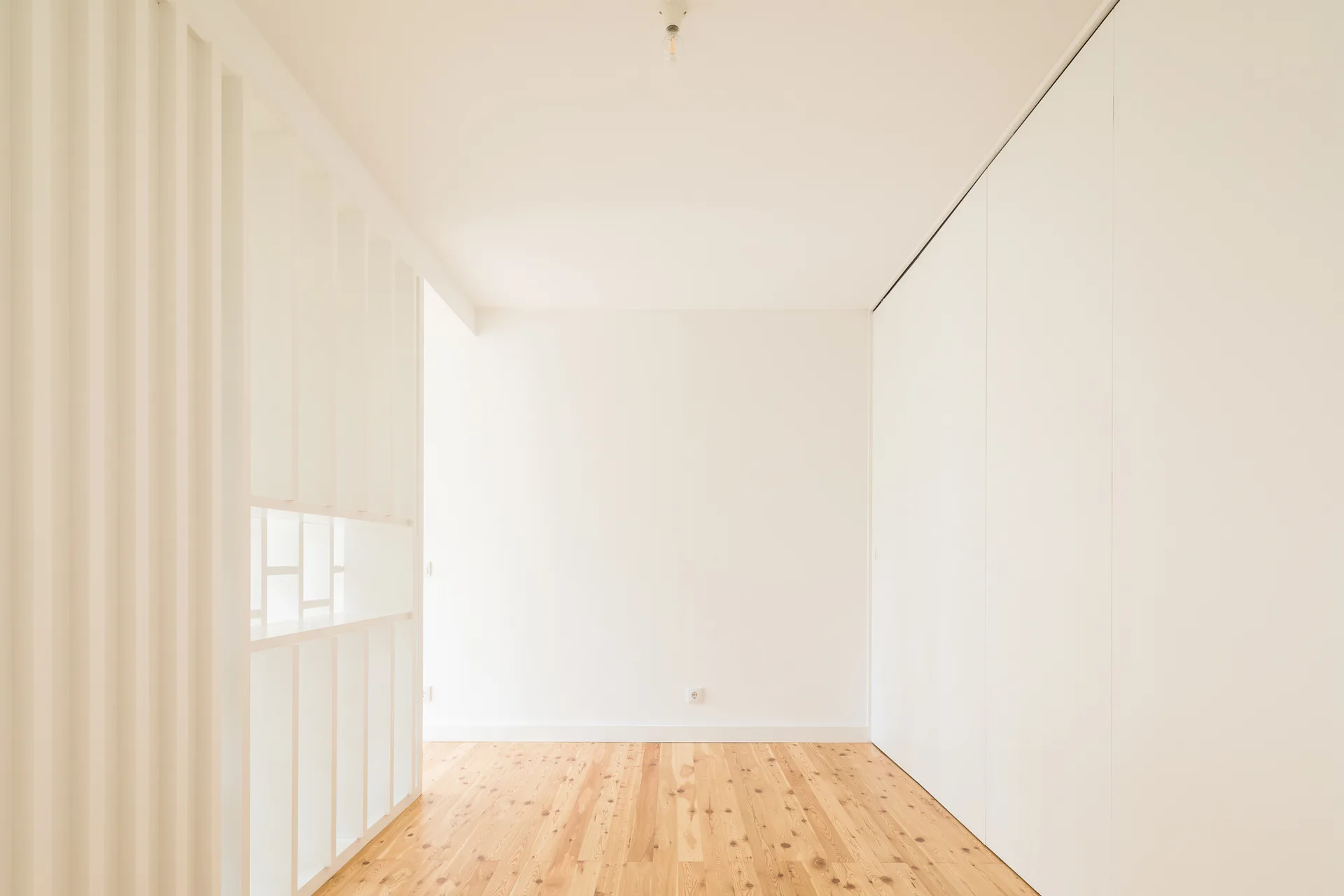 An empty minimalist room with light wood flooring, white walls, a vertical slatted divider, and floor-to-ceiling closet doors.