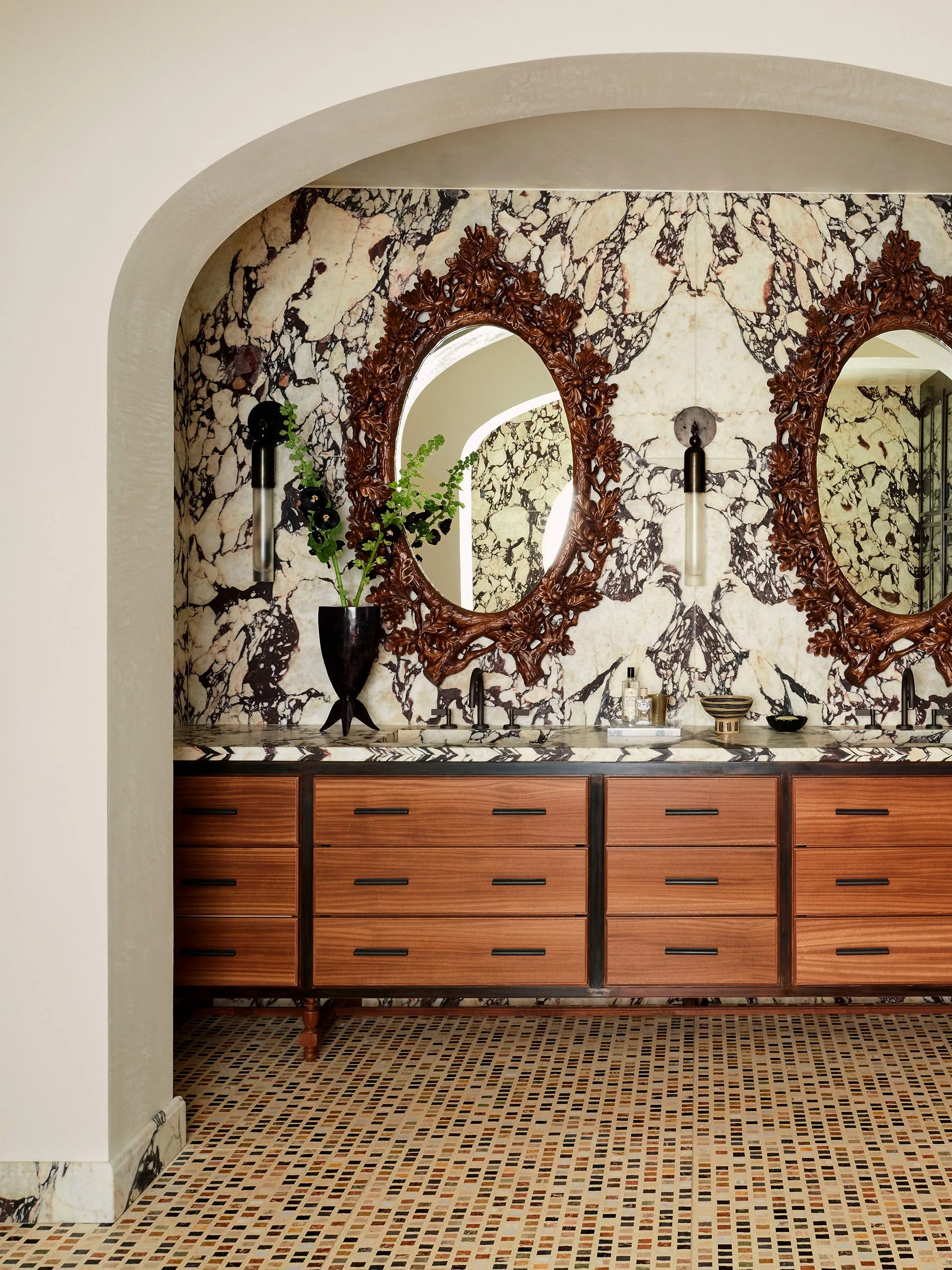 A bathroom double vanity with warm wood cabinets and ornate carved mirrors is set against a dramatically veined marble wall under a plaster archway.