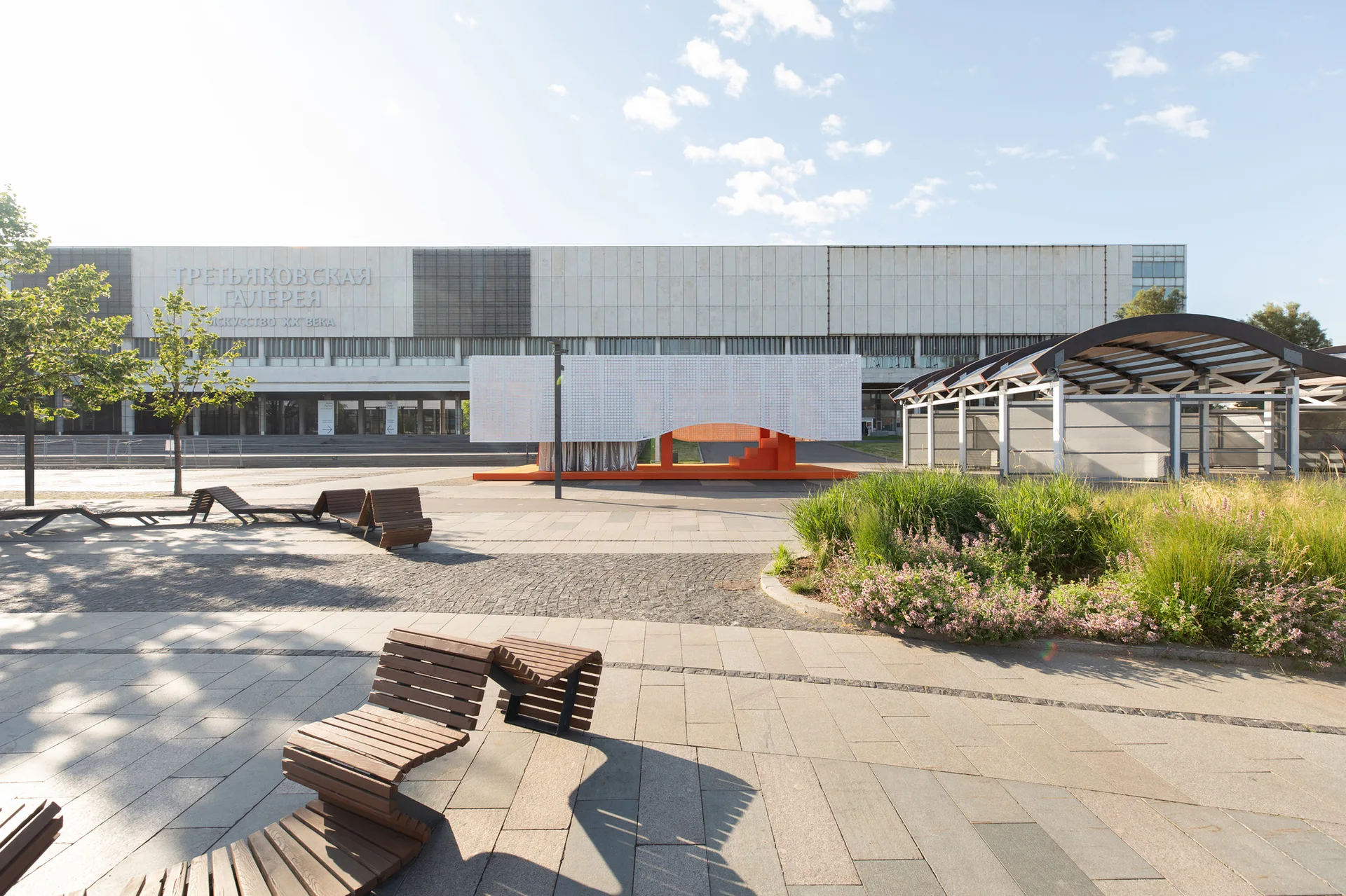 An outdoor paved public plaza featuring a modern white and orange geometric pavilion, wave-shaped slatted wooden benches, and landscaping.