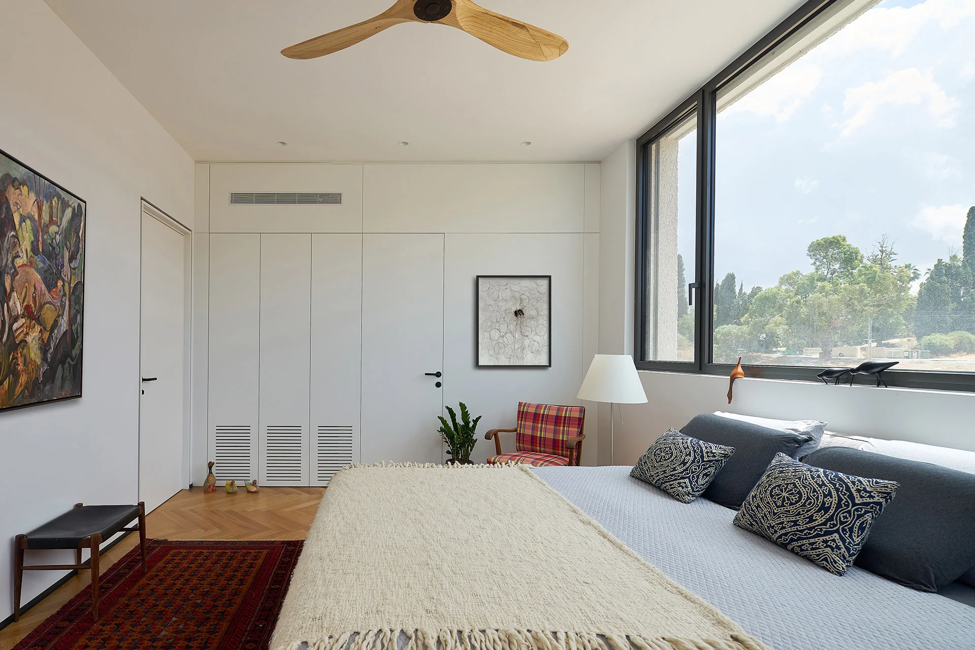 A minimalist bedroom featuring a bed with patterned pillows, white built-in wardrobes, a wood ceiling fan, and herringbone hardwood flooring.