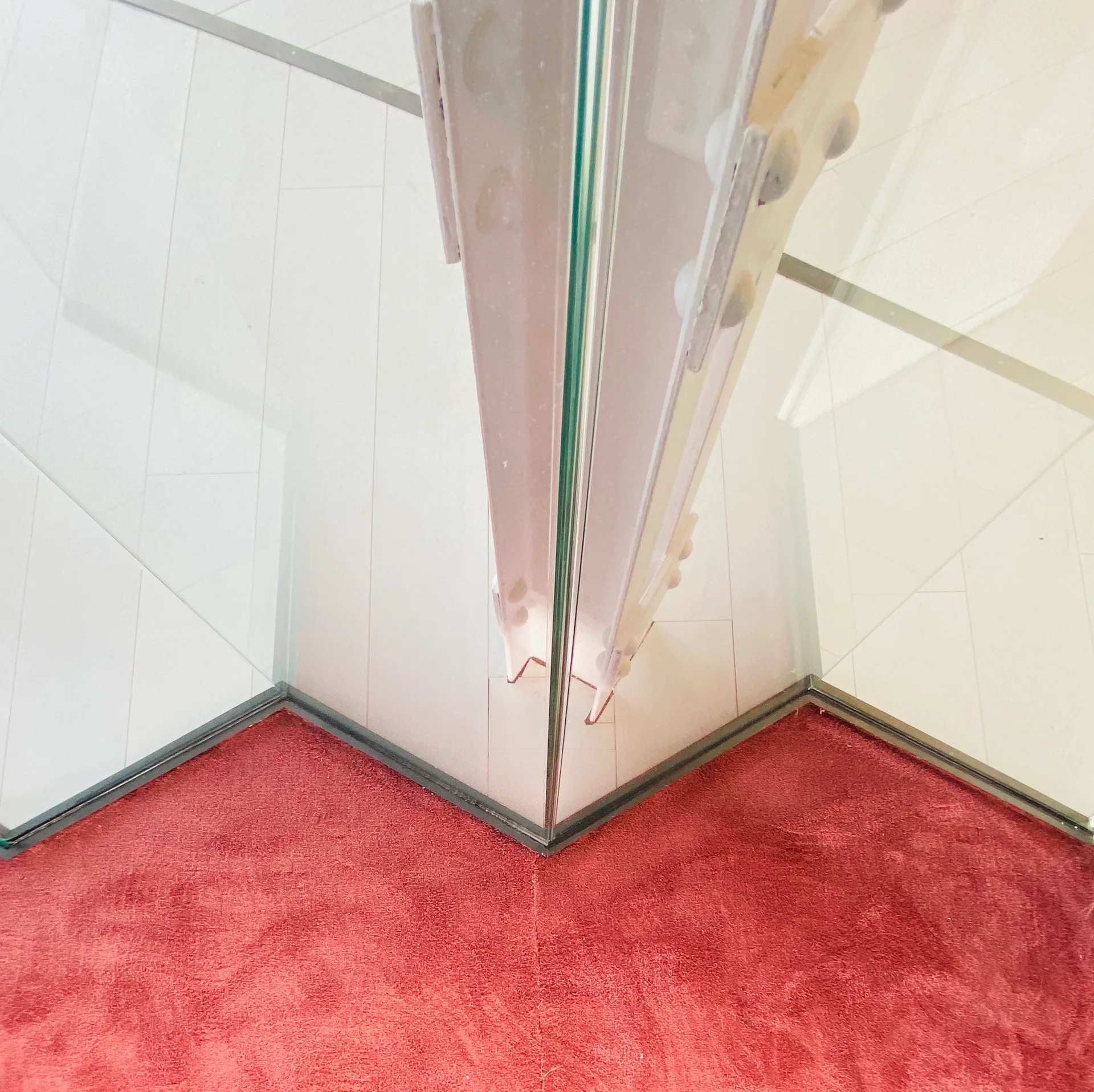 A close-up view of a clear glass partition corner meeting textured red carpet with vertical white wall paneling in the background.
