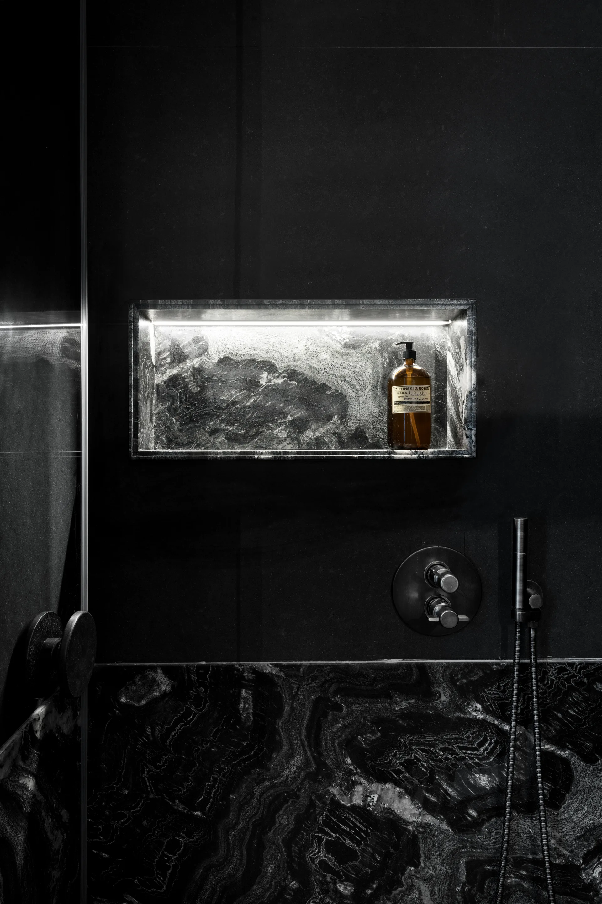 A dark stone shower wall features a recessed rectangular niche illuminated by an LED light strip above a metal faucet control.
