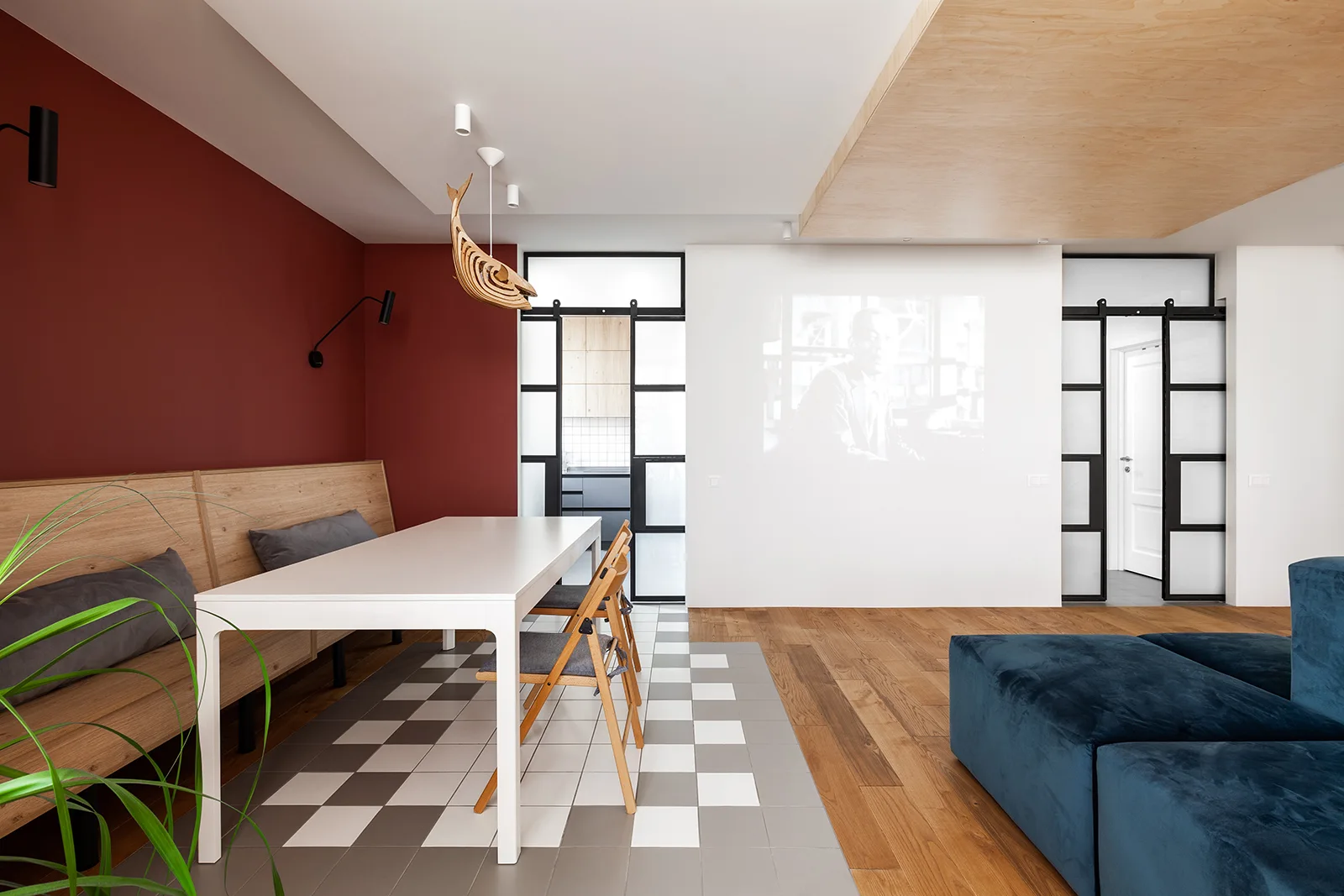 A modern dining area features a white table on a grey and white checkerboard tile floor, a red accent wall with a wooden bench, and black sliding doors.