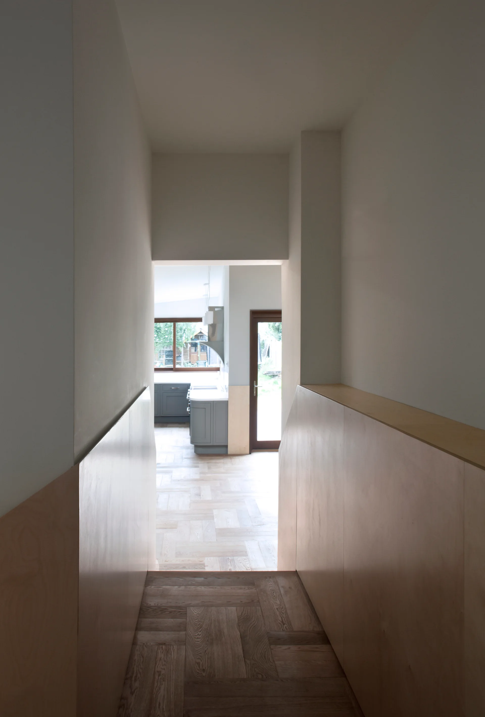 A view looking down a narrow hallway with light wood wall paneling and parquet floors toward a bright kitchen with grey cabinets.
