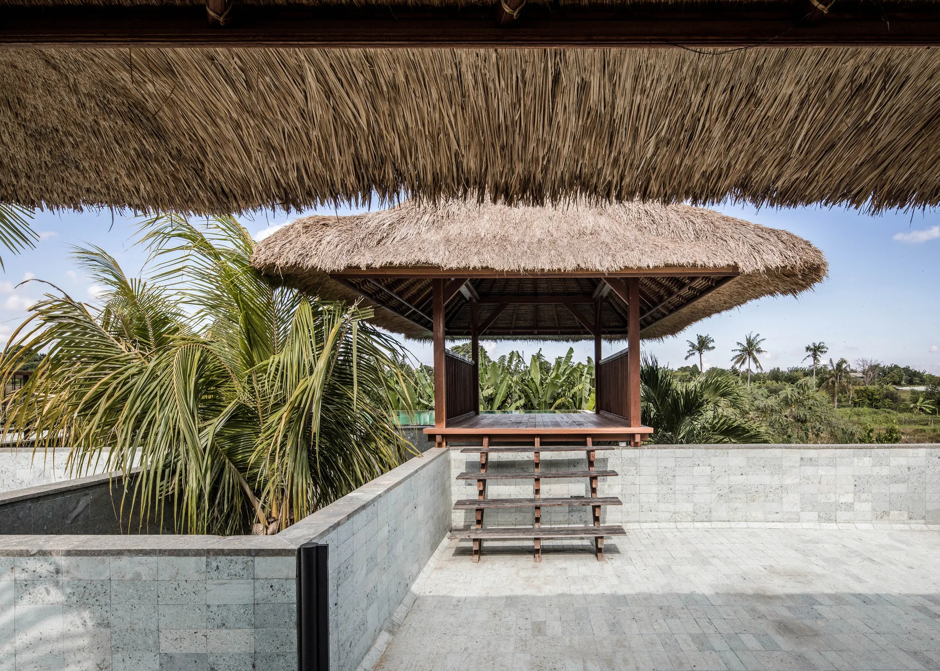 A raised wooden pavilion with a thatched roof sits on a grey stone-tiled terrace next to a lush green palm tree.
