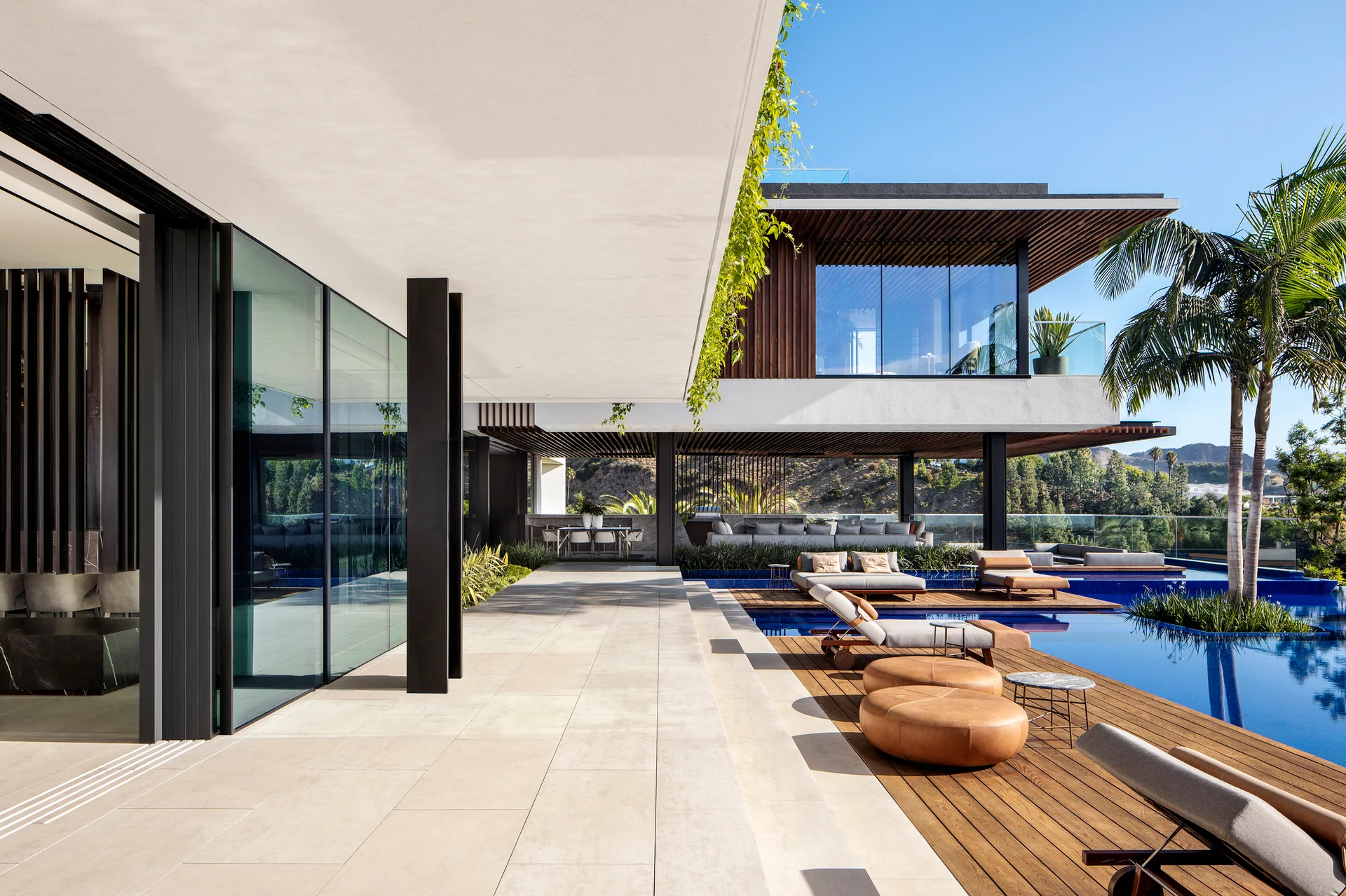 A modern outdoor patio features a blue swimming pool, wood decking with loungers, tan round ottomans, and a slatted timber overhang.