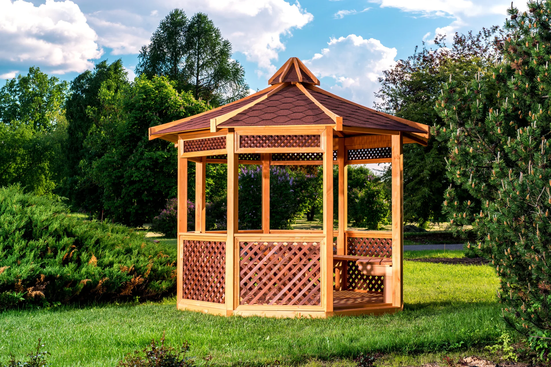 A hexagonal wooden gazebo with a brown shingled roof and diagonal lattice panels sits on a green lawn surrounded by trees.
