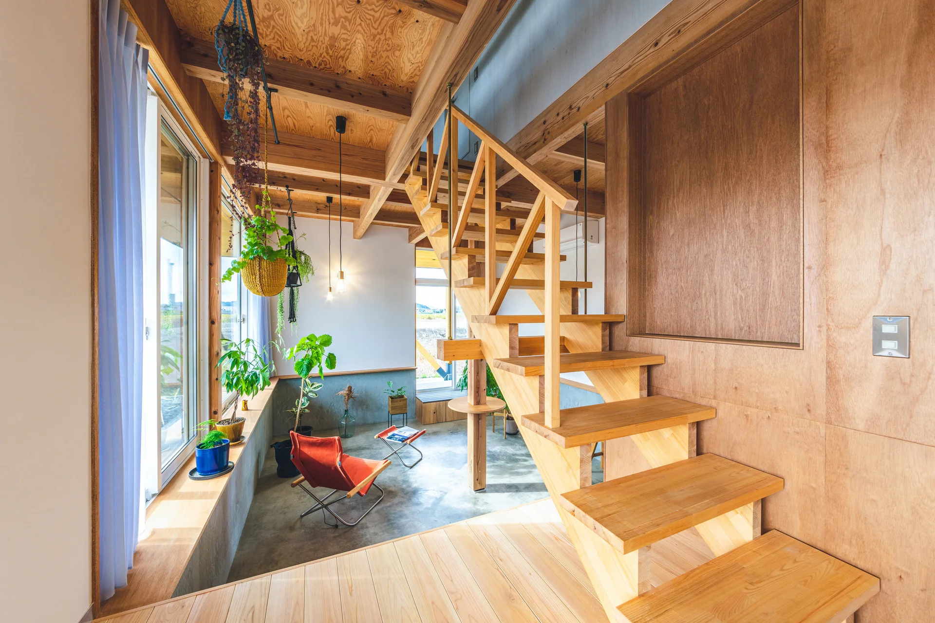 A minimalist living space featuring an open-riser wooden staircase, a concrete floor, plywood paneling, and an orange folding chair.