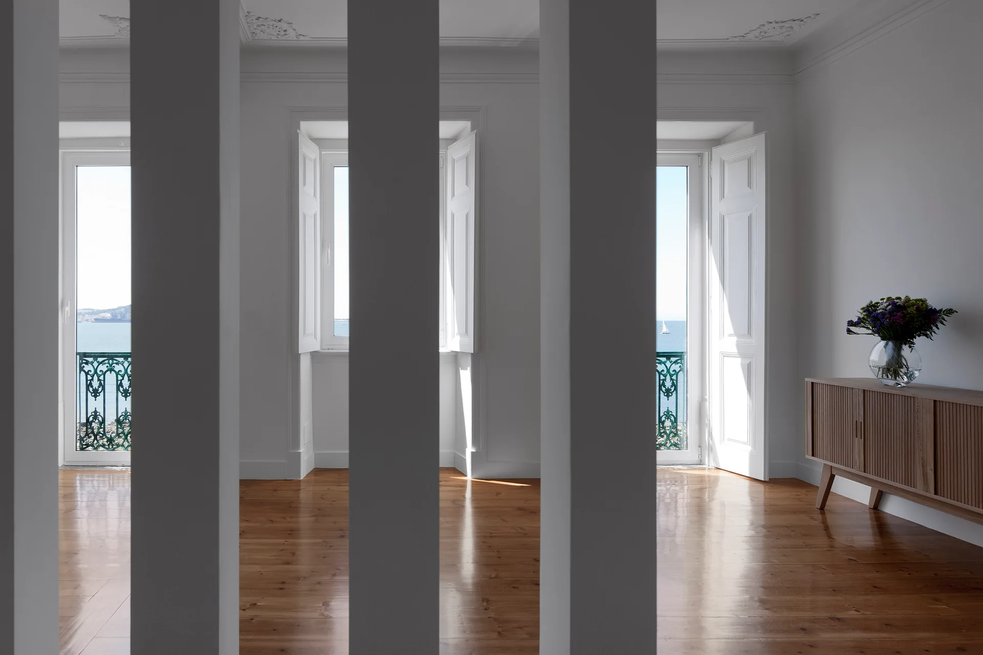 A bright white coastal room features glossy wood floors, a wooden console with a flower vase, and glass doors opening to a balcony with sea views.