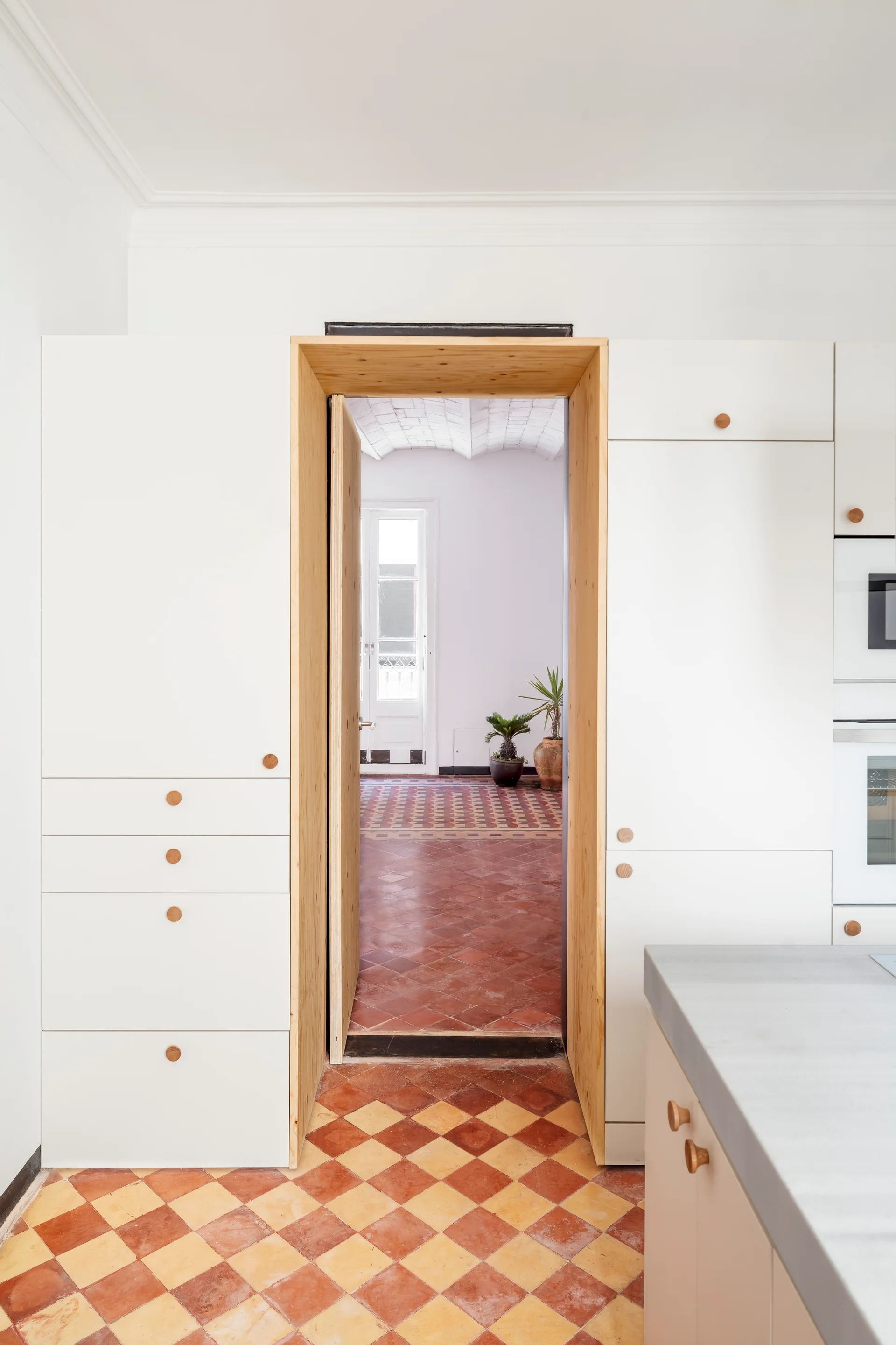 White built-in kitchen cabinets with round wood knobs flank a natural wood-trimmed doorway that opens to a room with terracotta checkerboard tile flooring.