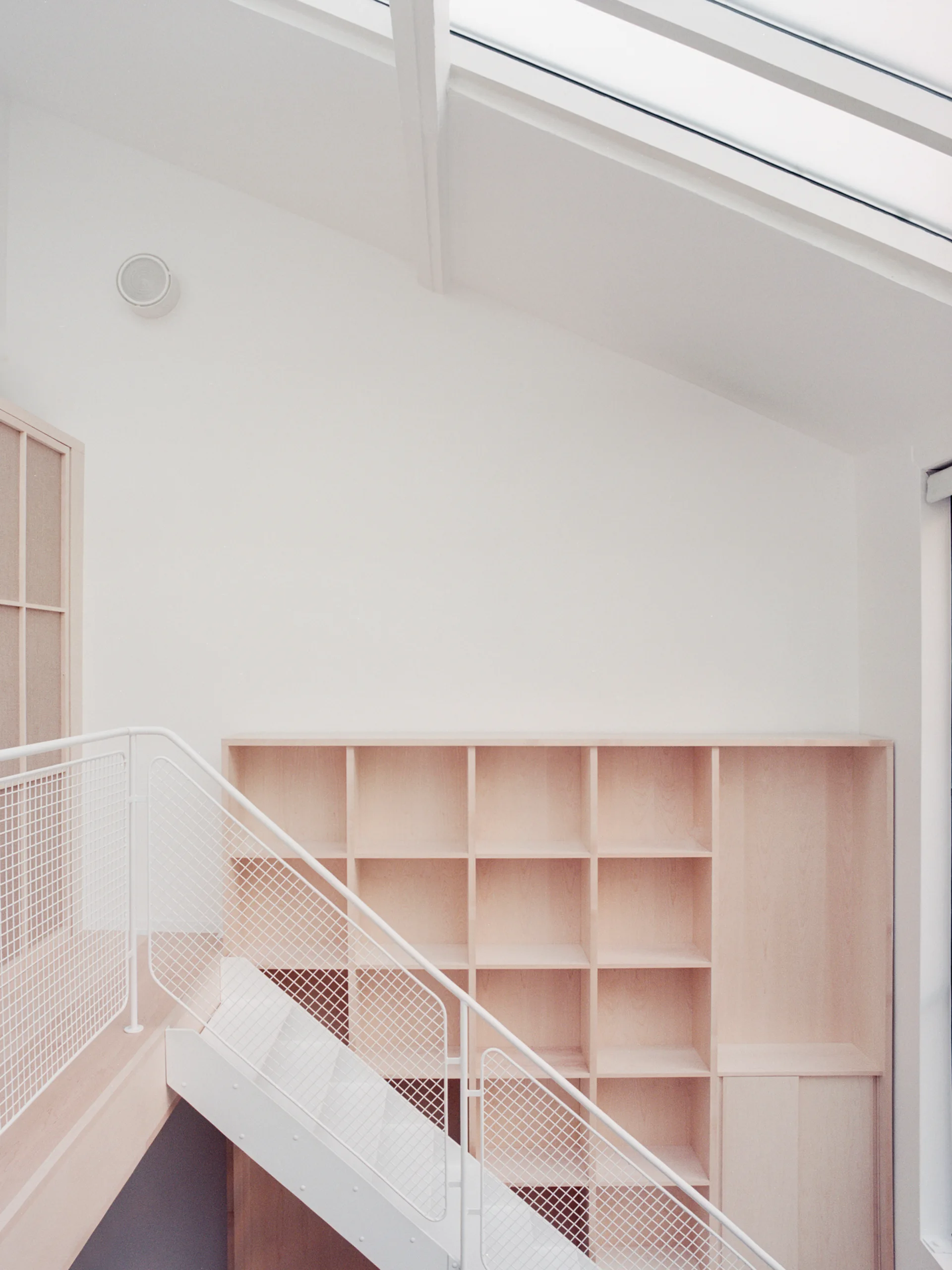 An interior staircase featuring a white wire mesh railing next to a light wood grid bookshelf under a skylight.