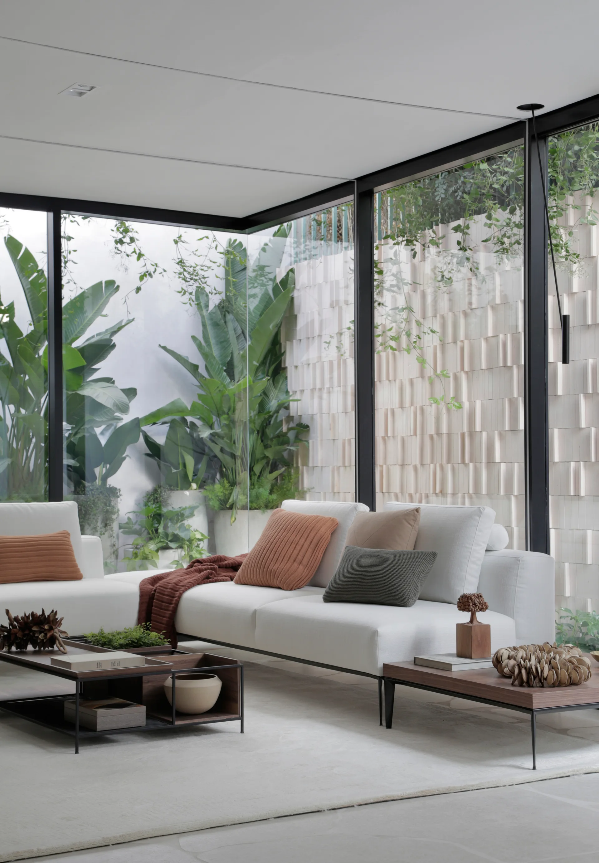A modern white sectional sofa with terracotta pillows sits beside large glass walls showcasing outdoor tropical plants.