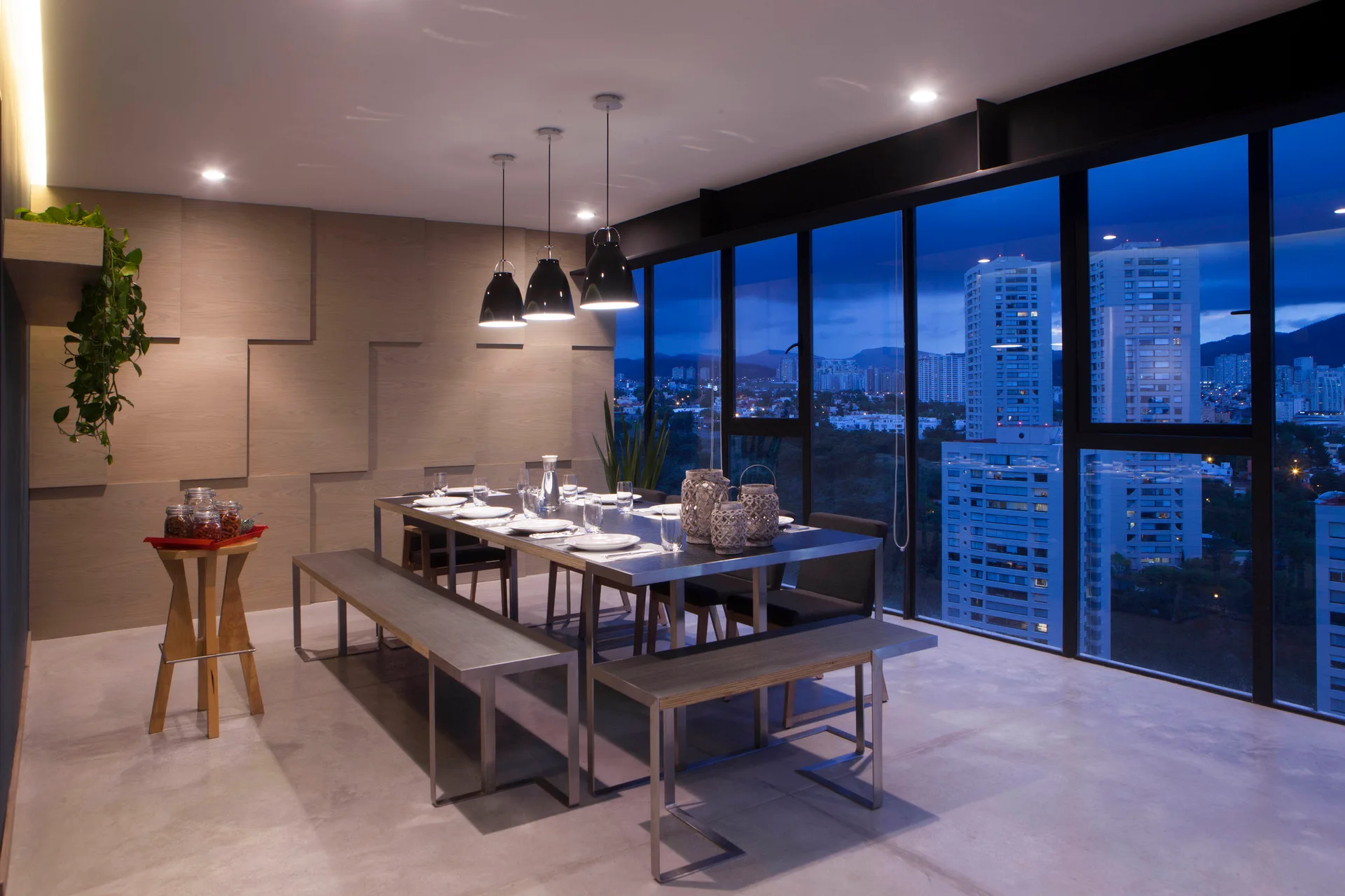 A modern dining room features a long table with benches, three black pendant lights, and a textured geometric wood wall overlooking a city at night.
