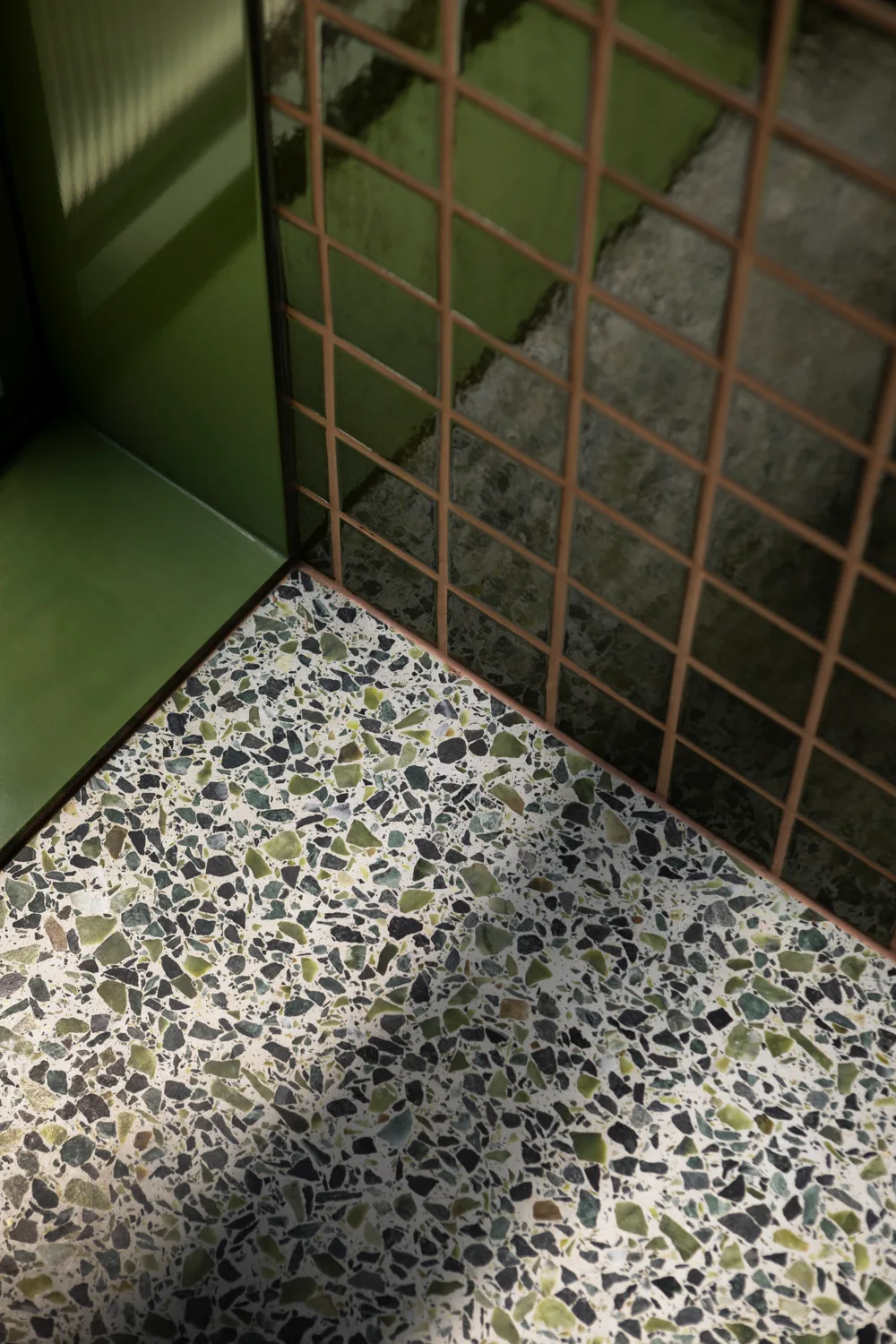 A close-up of a corner showcasing a speckled green terrazzo floor beneath a dark green grid tiled wall and a matte green solid panel.
